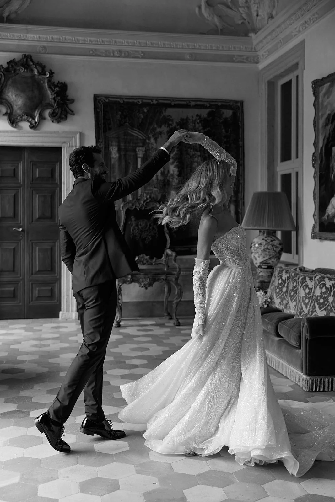 Ein Mann und eine Frau tanzen in einem eleganten Raum. Der Mann trägt einen schwarzen Anzug, die Frau trägt ein langes, schimmerndes Hochzeitskleid. Es scheint ein formeller Tanz, möglicherweise eine Hochzeit, zu sein.