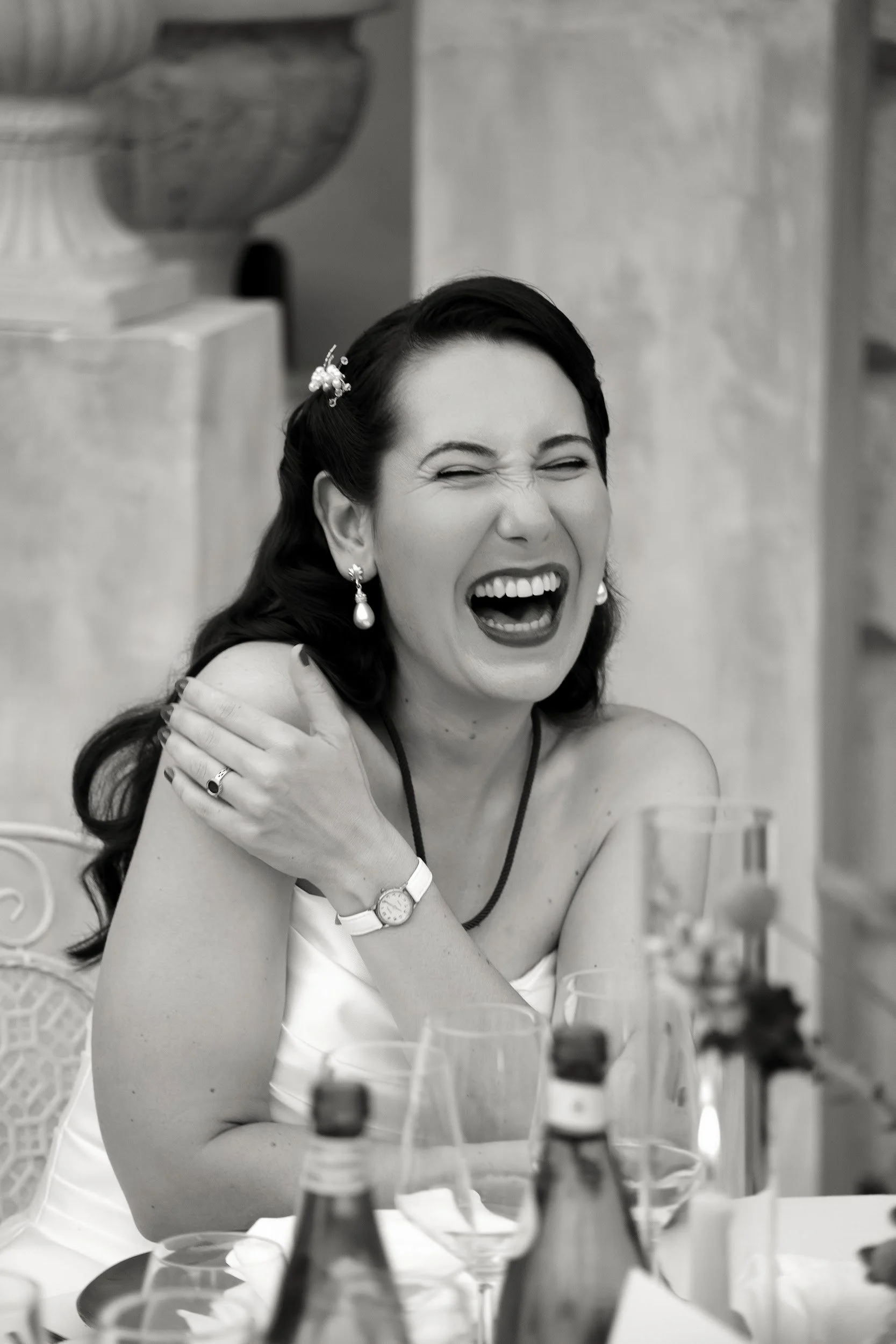 Frau lacht lachend auf einer Party, trägt elegante Kleidung, Schmuck und schaut fröhlich, im Hintergrund Dekoration und Tische.