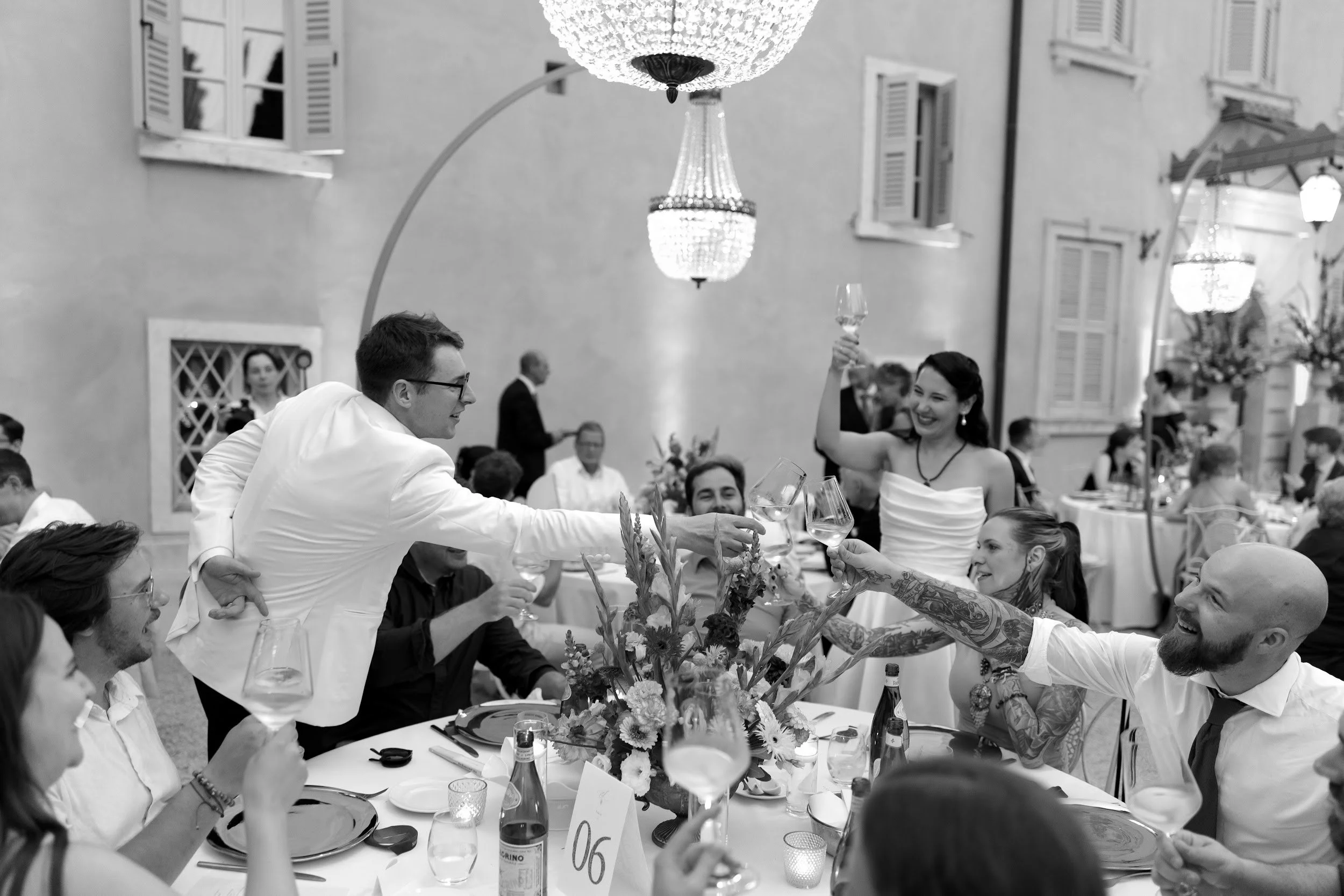 Bildausschnitt eines festlichen Abendessens bei einer Hochzeit, bei dem Menschen in eleganter Kleidung fröhlich an einem langen Tisch sitzen, umgeben von Blumen und Kerzen.