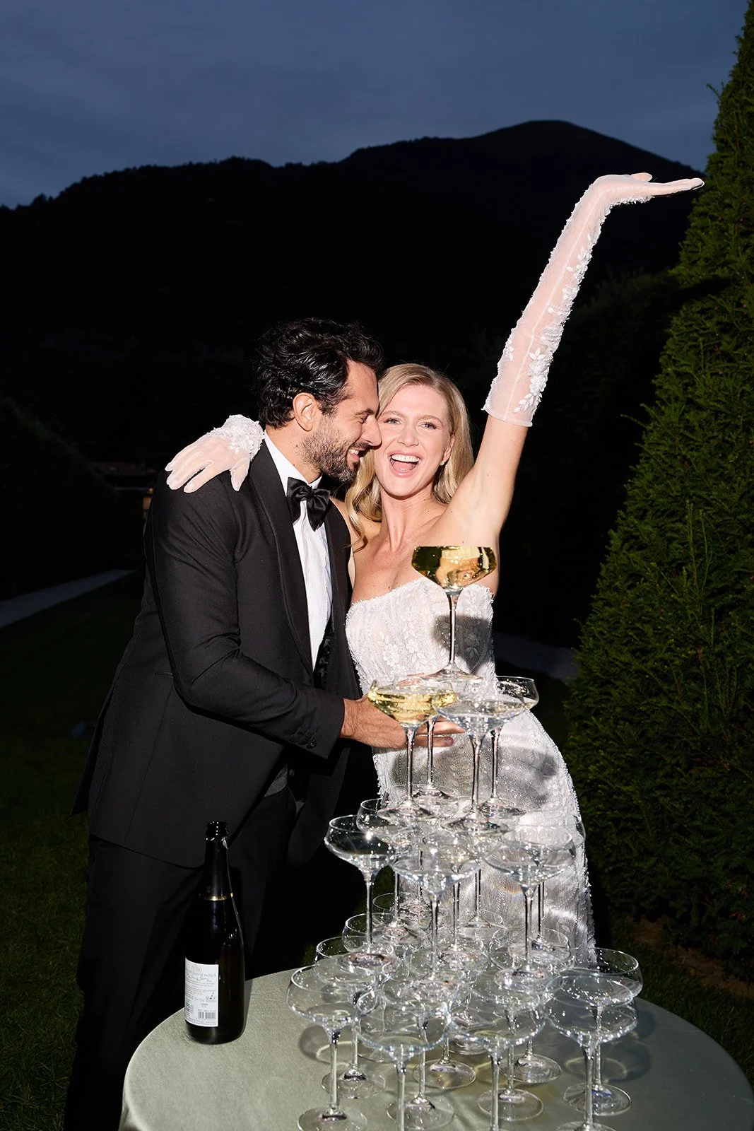 Ein glückliches Brautpaar beim Champagner-Anschnitt auf einer Hochzeit. Sie lachen, während sie einen Champagnerpyramide aus Gläsern aufbauen, im Hintergrund Berge und ein blauer Himmel bei Dämmerung.