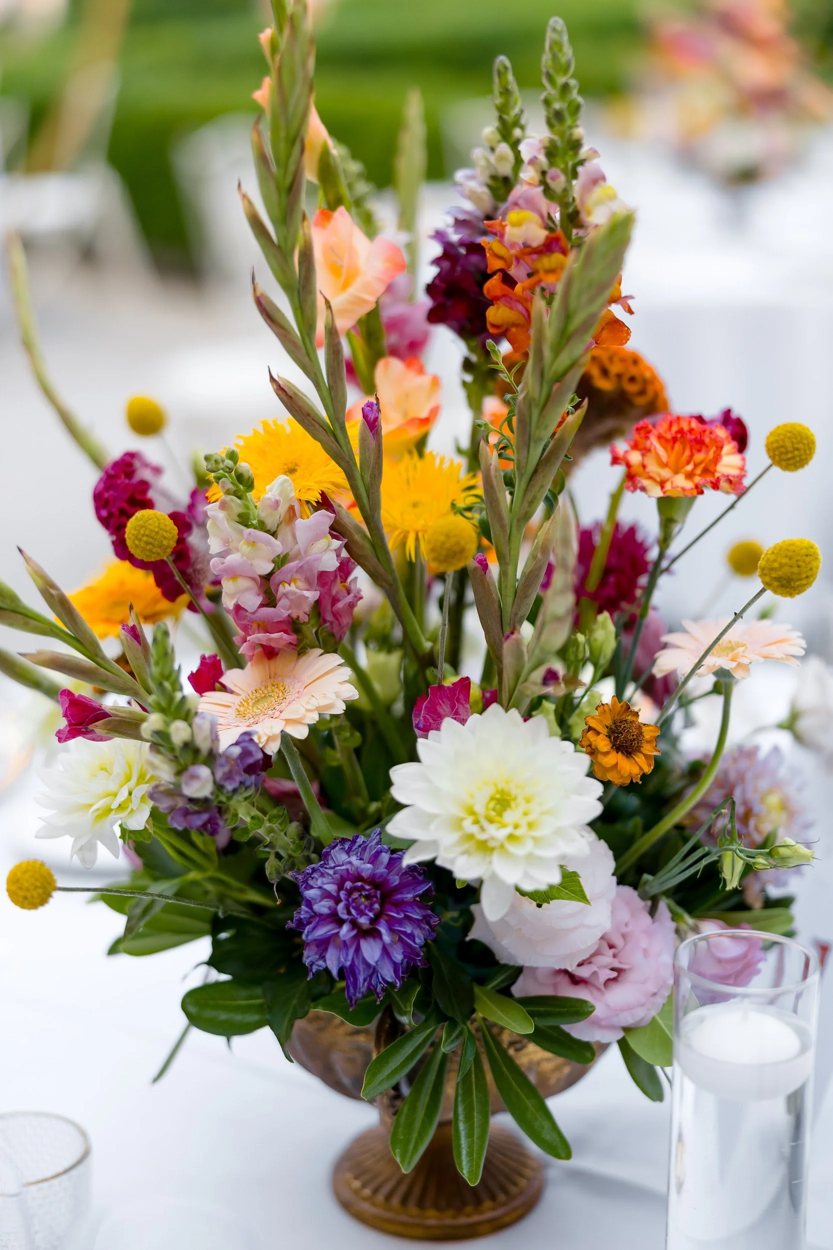 Bunte Blumenarrangement in einer Vase, umgeben von Gläsern auf einem weißen Tisch.