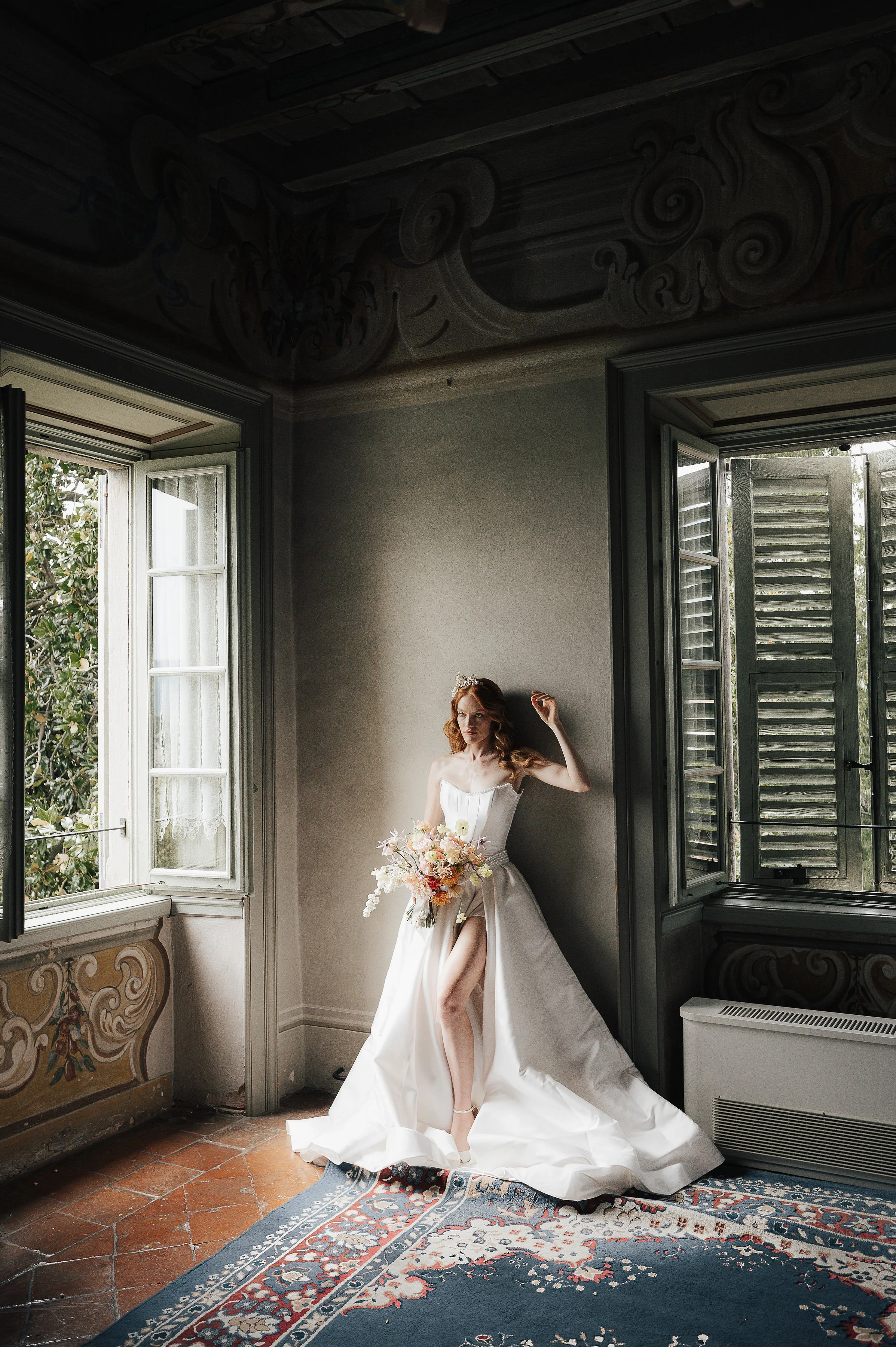 Eine junge Frau in einem weißen Brautkleid steht in einem altmodischen Raum mit offenen Fenstern, die viel natürliches Licht hereinlassen. Sie hält einen großen Blumenstrauß und posiert elegant auf einem Teppich auf dem Boden.