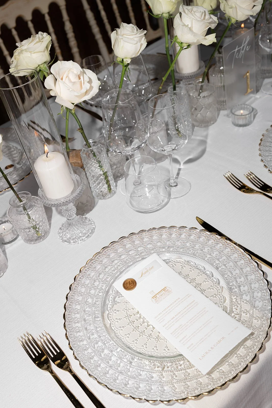 Eleganter gedeckter Tisch mit weißen Rosen, Kerzen, Kristallglas, goldene Besteck, elegantes Menükarten und weißen Platzdeckchen.