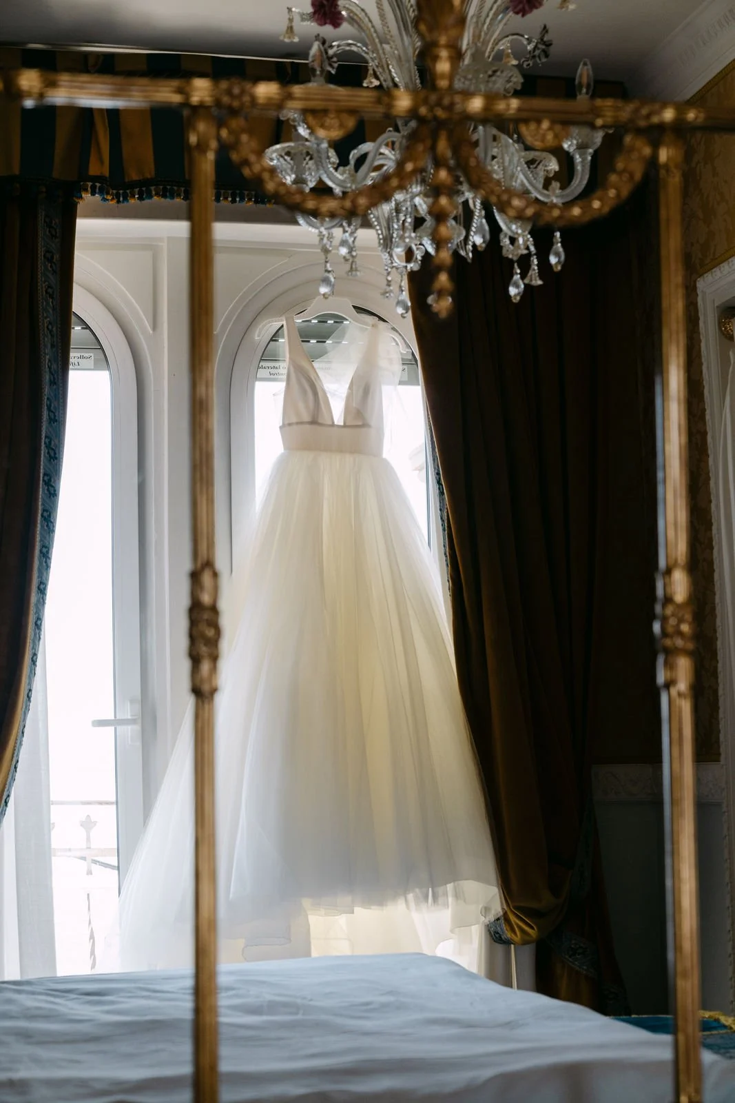 Elegantes weißes Brautkleid hängt in einem Raum vor einem Fenster, sichtbar durch eine goldene Ladentür mit Kristallkronleuchter im Vordergrund.