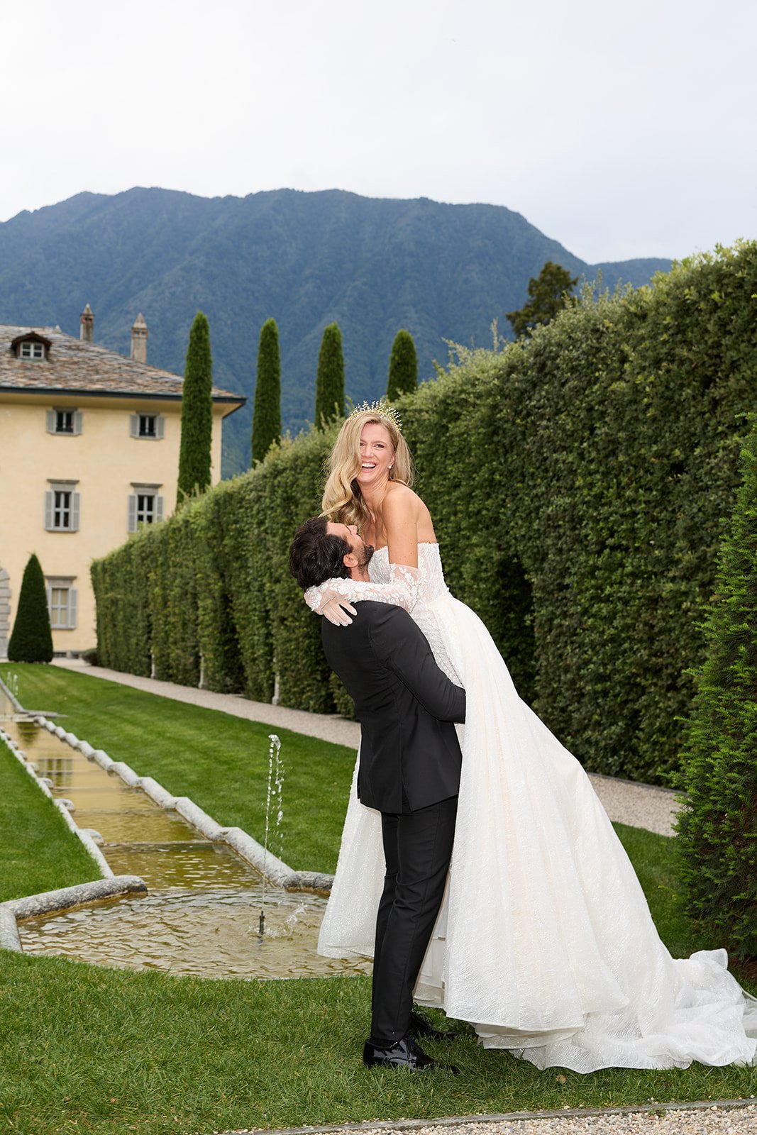 Ein Brautpaar bei einer Hochzeit im Freien, der Bräutigam hebt die Braut hoch, umgeben von gepflegtem Garten, hohen Bäumen und Bergen im Hintergrund.