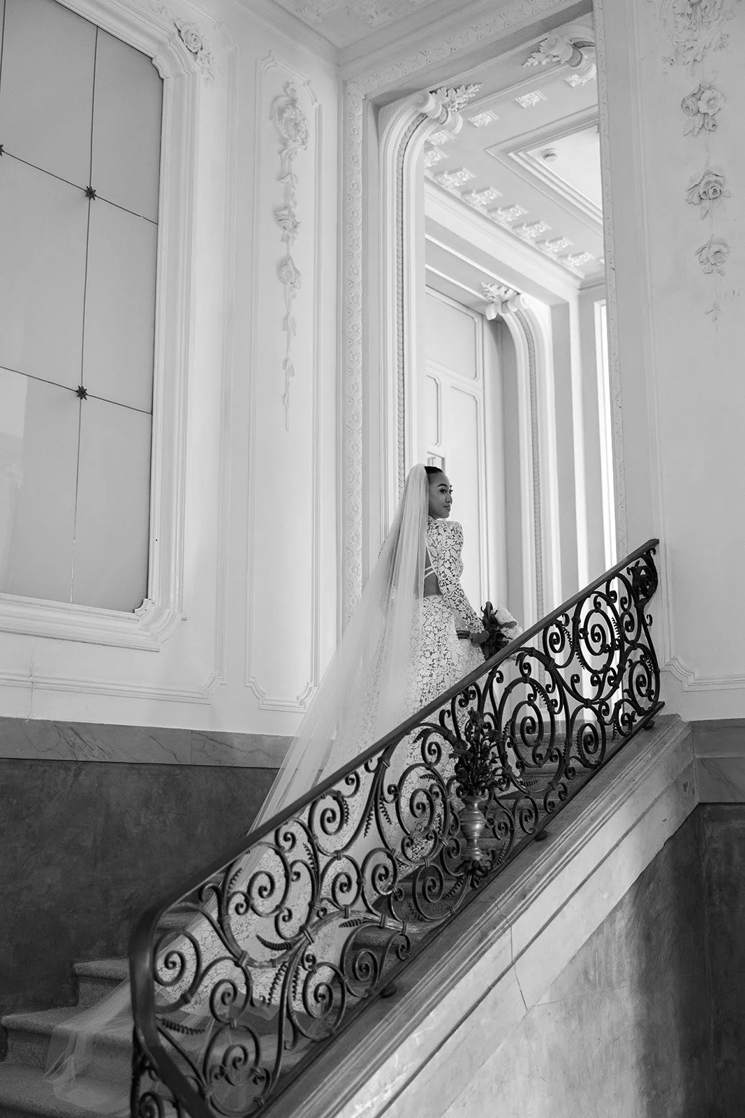 Schwarz-weiße Fotografie einer Braut in einem eleganten Hochzeitskleid auf einer Treppe mit kunstvoller Eisengeländer, in einem prunkvollen, mit Ornamenten verzierten Raum.