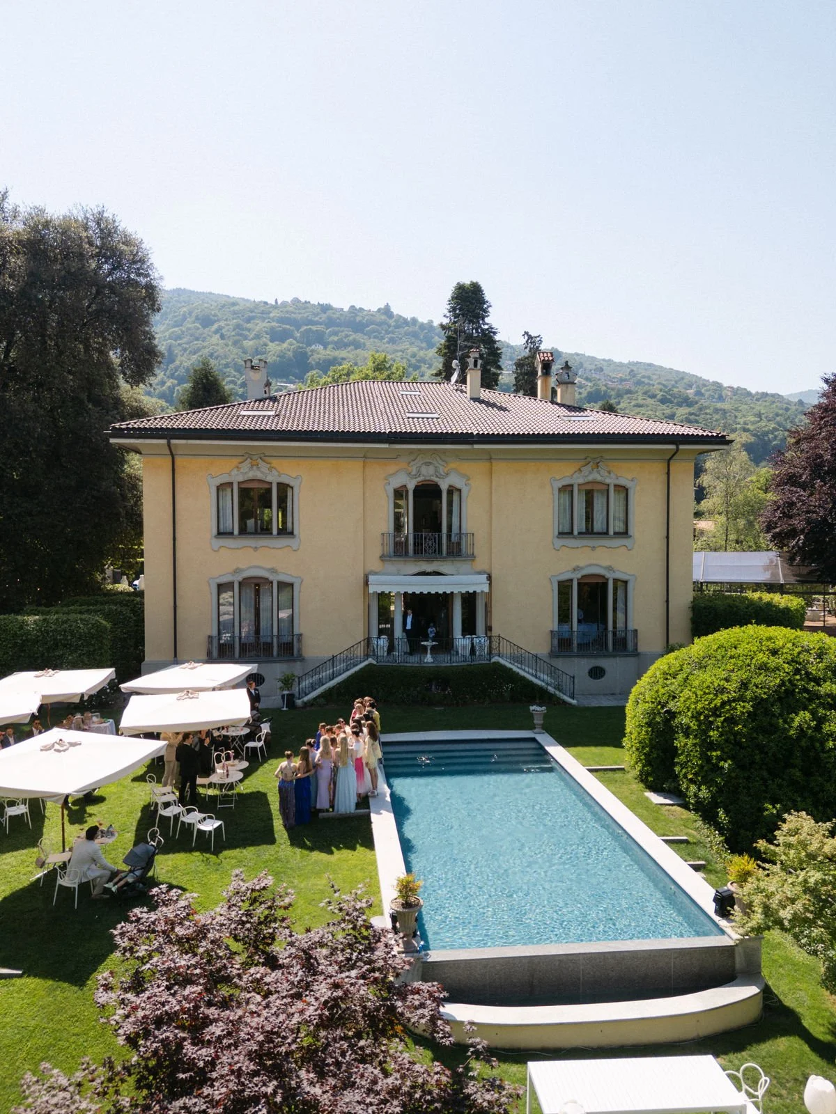 Ein mehrstöckiges, gelbes Haus mit Balkon und rotem Dach, umgeben von grünen Bäumen und Hügeln im Hintergrund. Im Vordergrund ein Schwimmbecken, um das sich eine Gruppe von Menschen in festlicher Kleidung versammelt, sowie Tische mit weißen Sonnensch