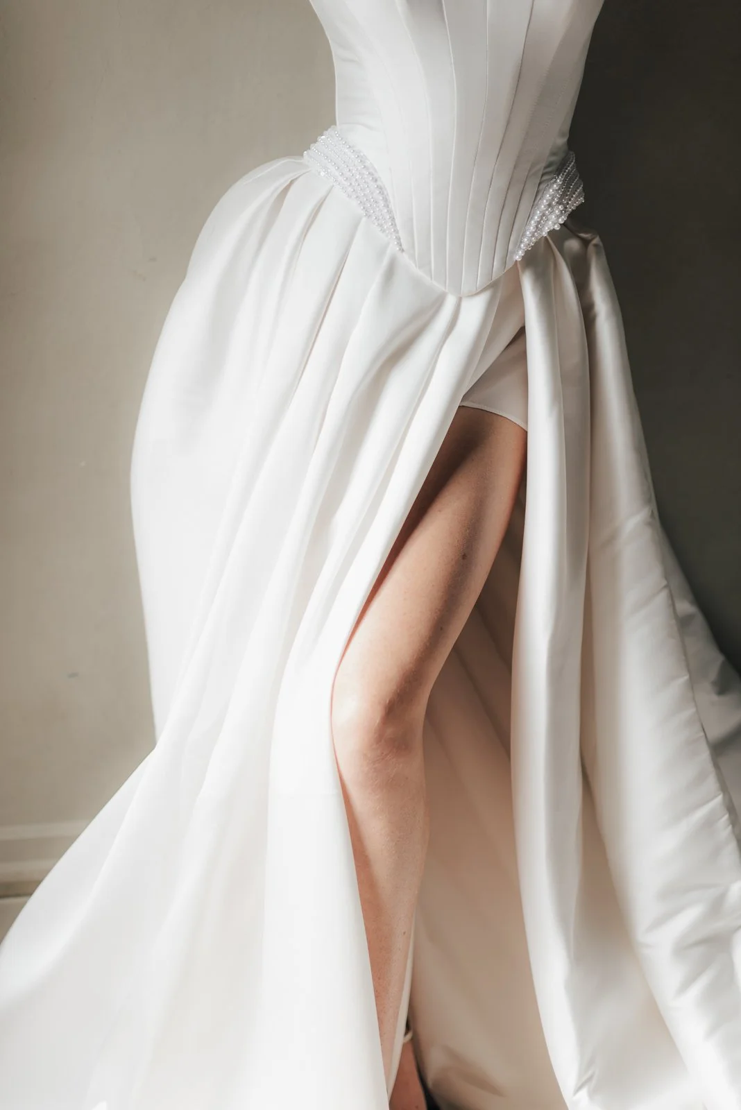 Nahaufnahme eines weißen Hochzeitskleids mit gerafften Stoffen, Perlenverzierungen und hoher Schlitze, die die Beine einer Person zeigen