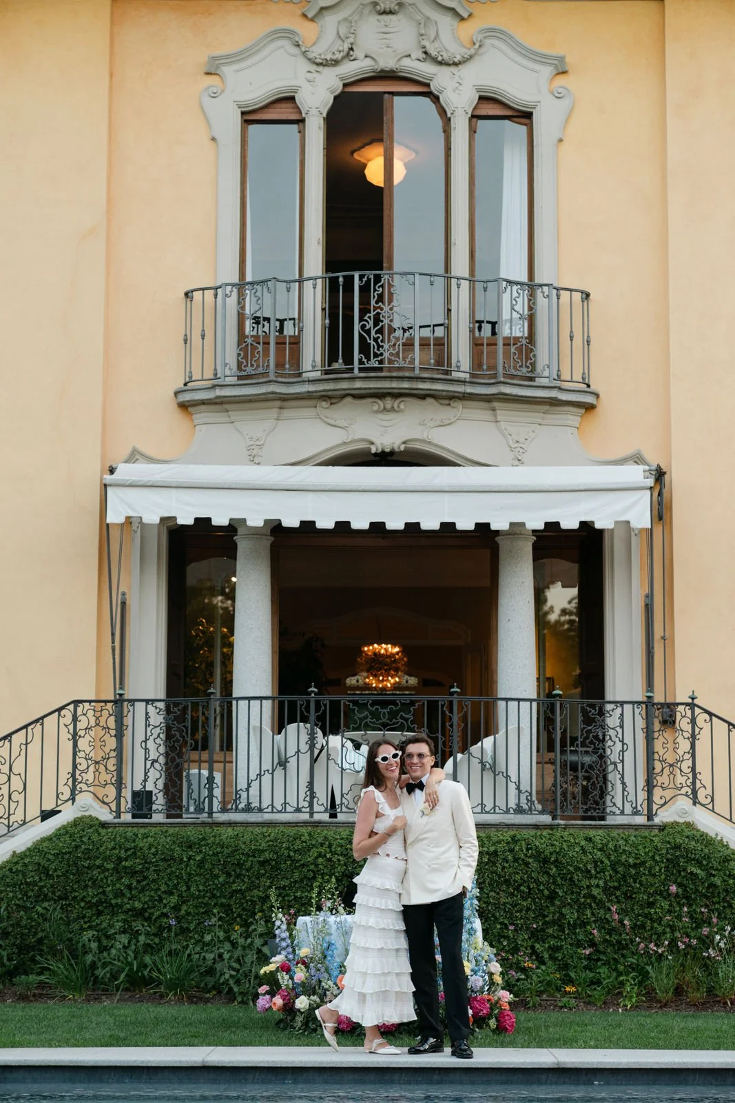 Ein Paar in Hochzeitskleidung steht vor einem mehrstöckigen historischen Gebäude mit Balkon, Blumenschmuck und einer Beleuchtung im Inneren.