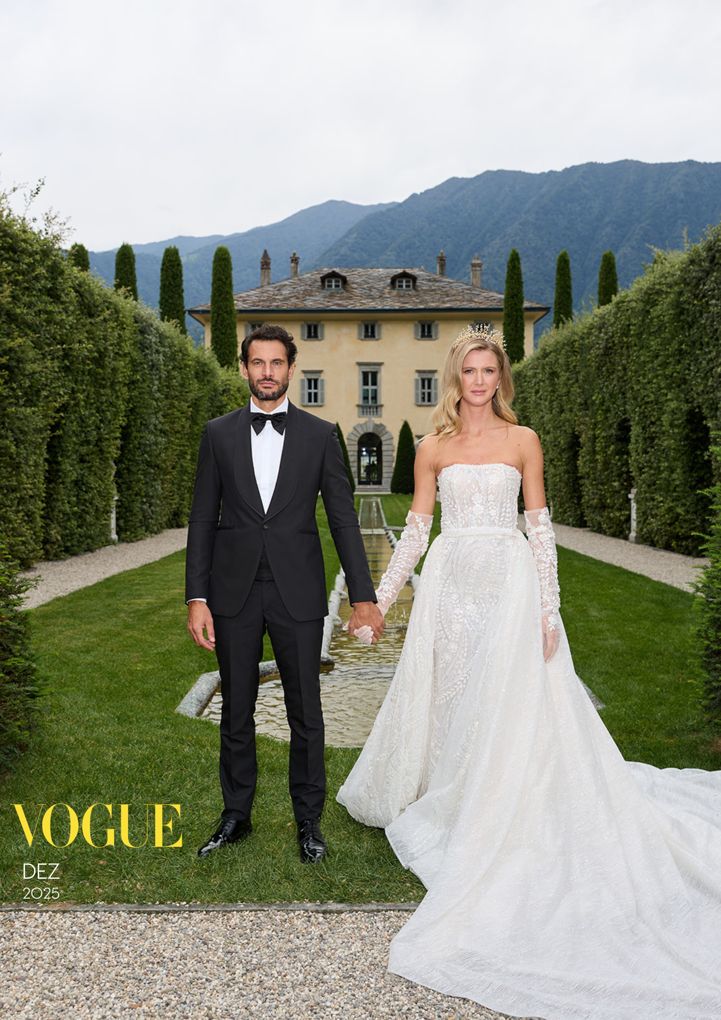 Ein Mann in einem schwarzen Anzug mit Fliege hält die Hand einer Frau in einem weißen Brautkleid mit Tiara, in einer grünen Gartenumgebung vor einem großen Haus mit Bergen im Hintergrund.