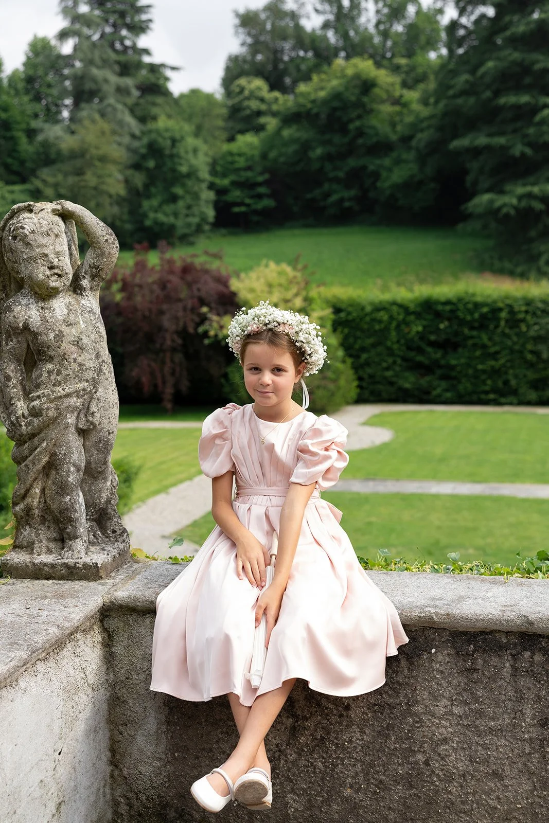 Ein junges Mädchen in einem rosa Kleid mit Blumenkranz sitzt auf einer Steinmauer neben einer Skulptur in einem Garten mit grünen Bäumen und Rasen.