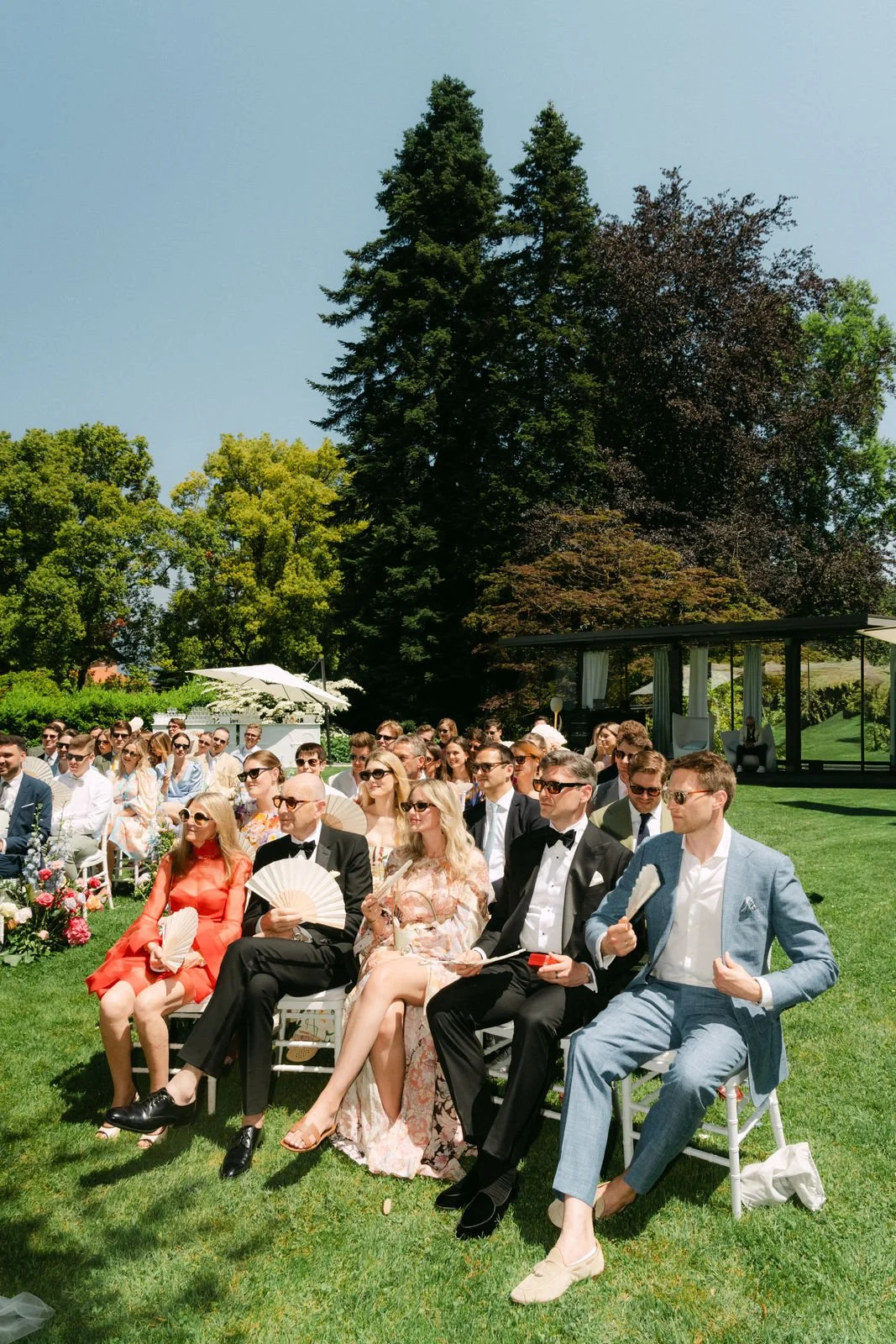 Herrschaften in formeller Kleidung bei einer Outdoor-Hochzeit, sitzend auf weißen Stühlen auf einer grünen Wiese, mit Bäumen im Hintergrund.