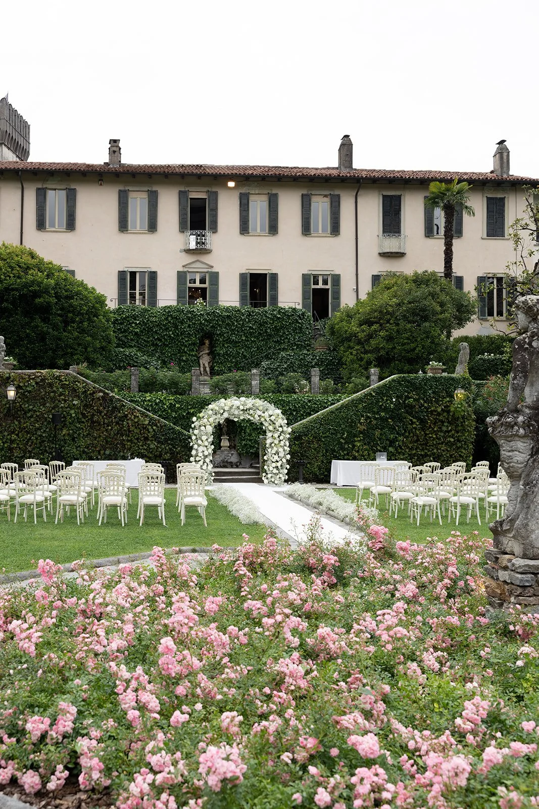 Ein Garten mit Stuhlreihen und Blumen, die für eine Hochzeit vorbereitet sind, vor einem alten Haus mit mehreren Fenstern.