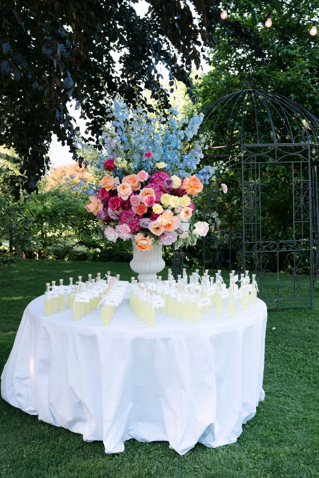 Ein runder Tisch mit weißen Tüchern, auf dem sich eine große Blumenvase mit bunten Blumen befindet. Vor der Vase sind kleine Flaschen mit Zweige, vermutlich für eine Hochzeitszeremonie.
