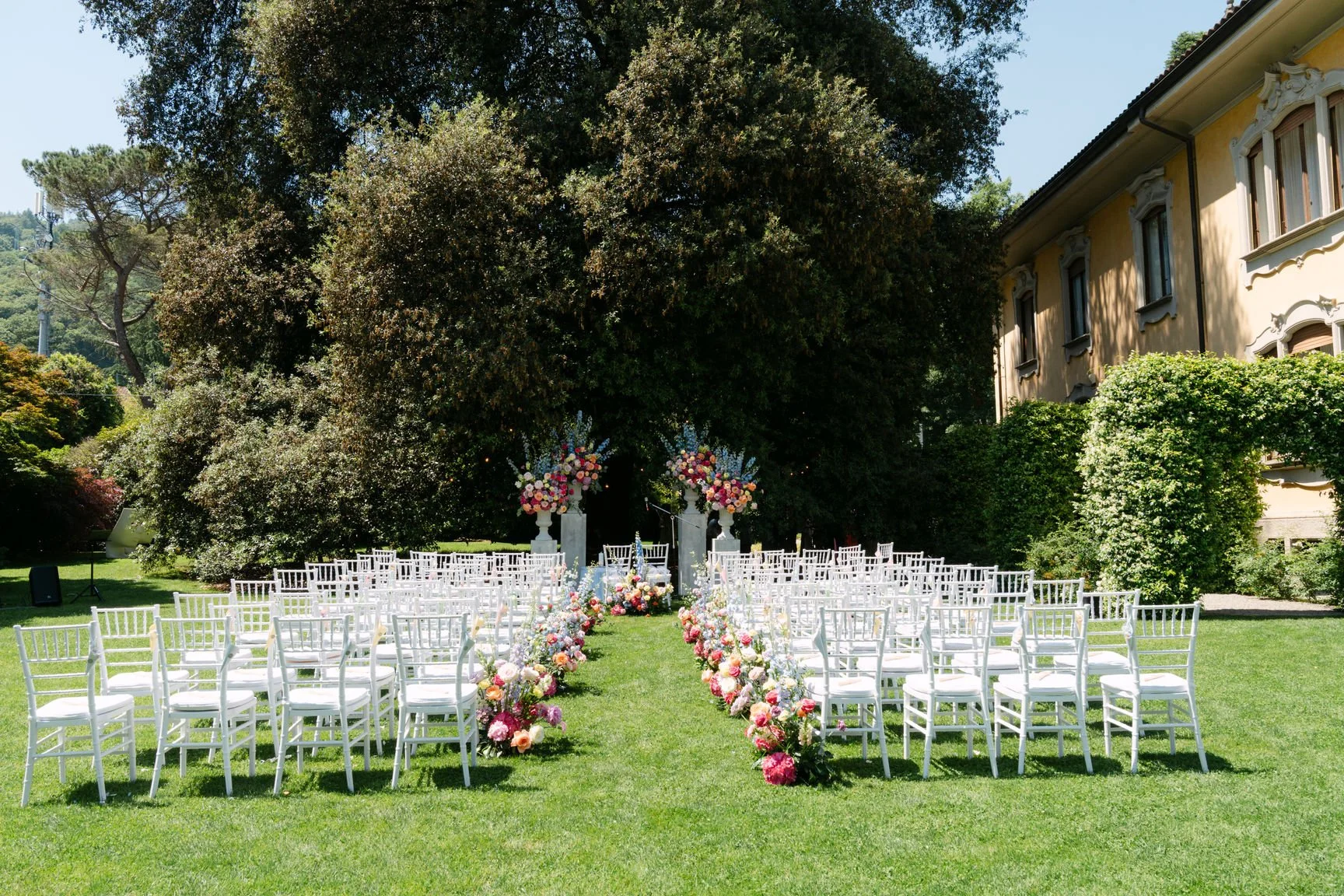 Eine wunderschöne Garten-Arrangements für eine Hochzeitszeremonie mit weißen Stühlen, Blumenarrangements und einer Altarwand, umgeben von Bäumen vor einem historischen Haus.