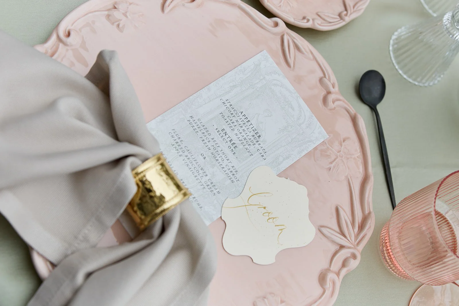 Ein Tisch mit rosa und pastellfarbenen Gegenständen, darunter eine Details eines Menüs, eine Kerze, eine Sektflöte und eine pinkfarbene Servierplatte, sowie eine elegant dekorierte Tischdekoration für eine Feier oder einen besonderen Anlass.