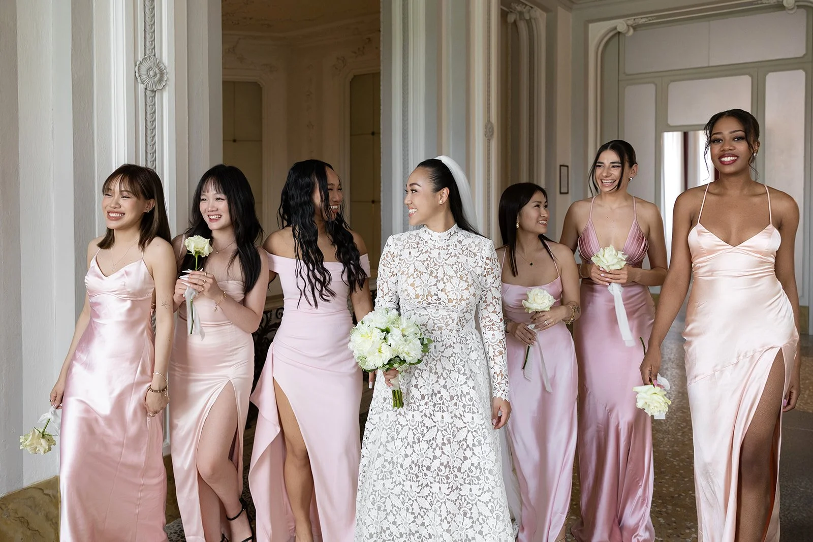 Eine Gruppe von Frauen in eleganten Kleidern, vermutlich bei einer Hochzeitsfeier, im Raum eines Saals, lachen und tragen Blumen.