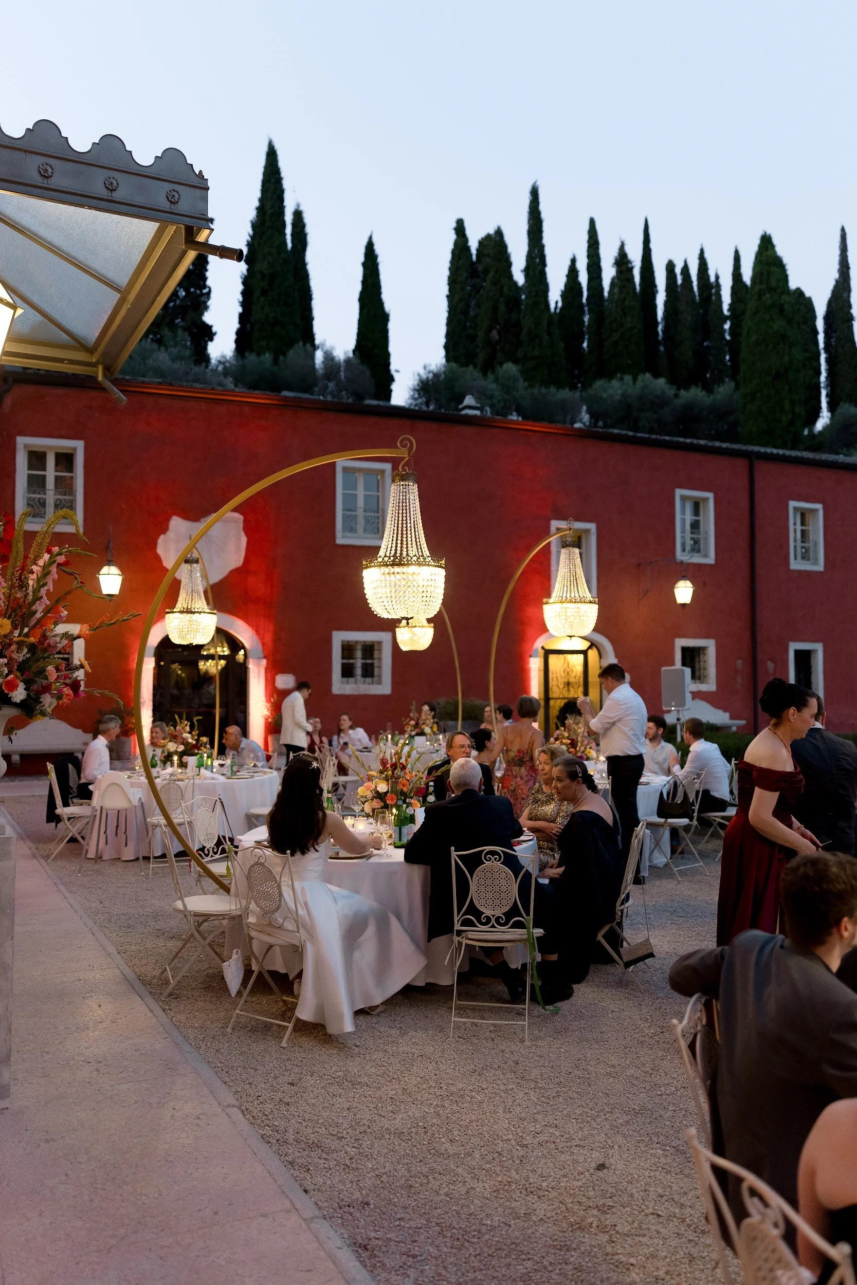 Ein festliches Abendessen im Freien mit mehreren Tischen, die mit weißen Tischtüchern und Blumenarrangements gedeckt sind. Es sind viele Gäste im eleganten Outfit, die sich unter großen, eleganten Kronleuchtern im Außenbereich eines roten Gebäudes ve