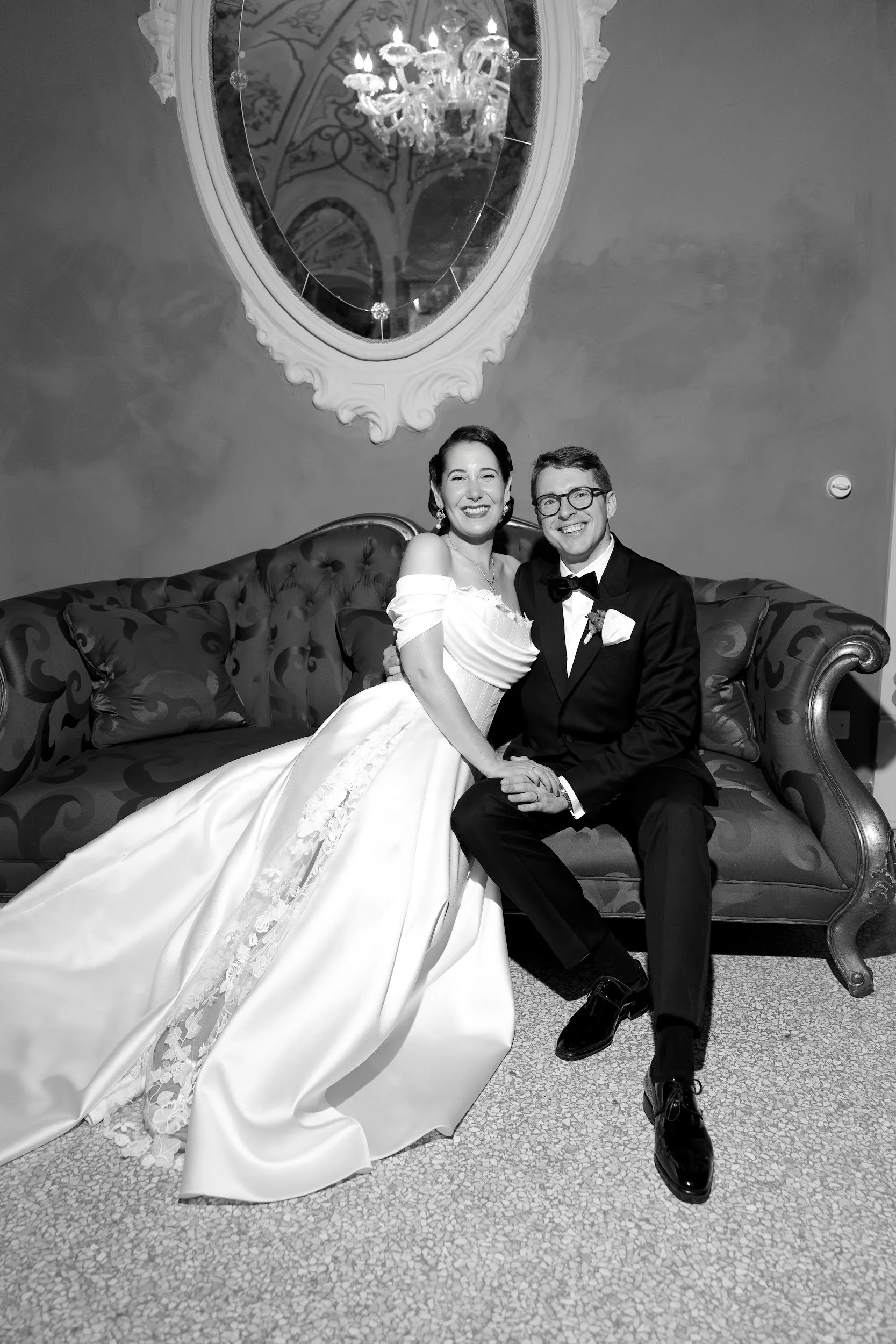 Ein braun-schwarzes Sofa mit einem Brautpaar in formeller Kleidung, die Braut trägt ein weißes Hochzeitskleid und der Bräutigam einen schwarzen Anzug mit Fliege. Hinten befindet sich ein großer antiker Spiegel und eine elegante Kronleuchterlampe.