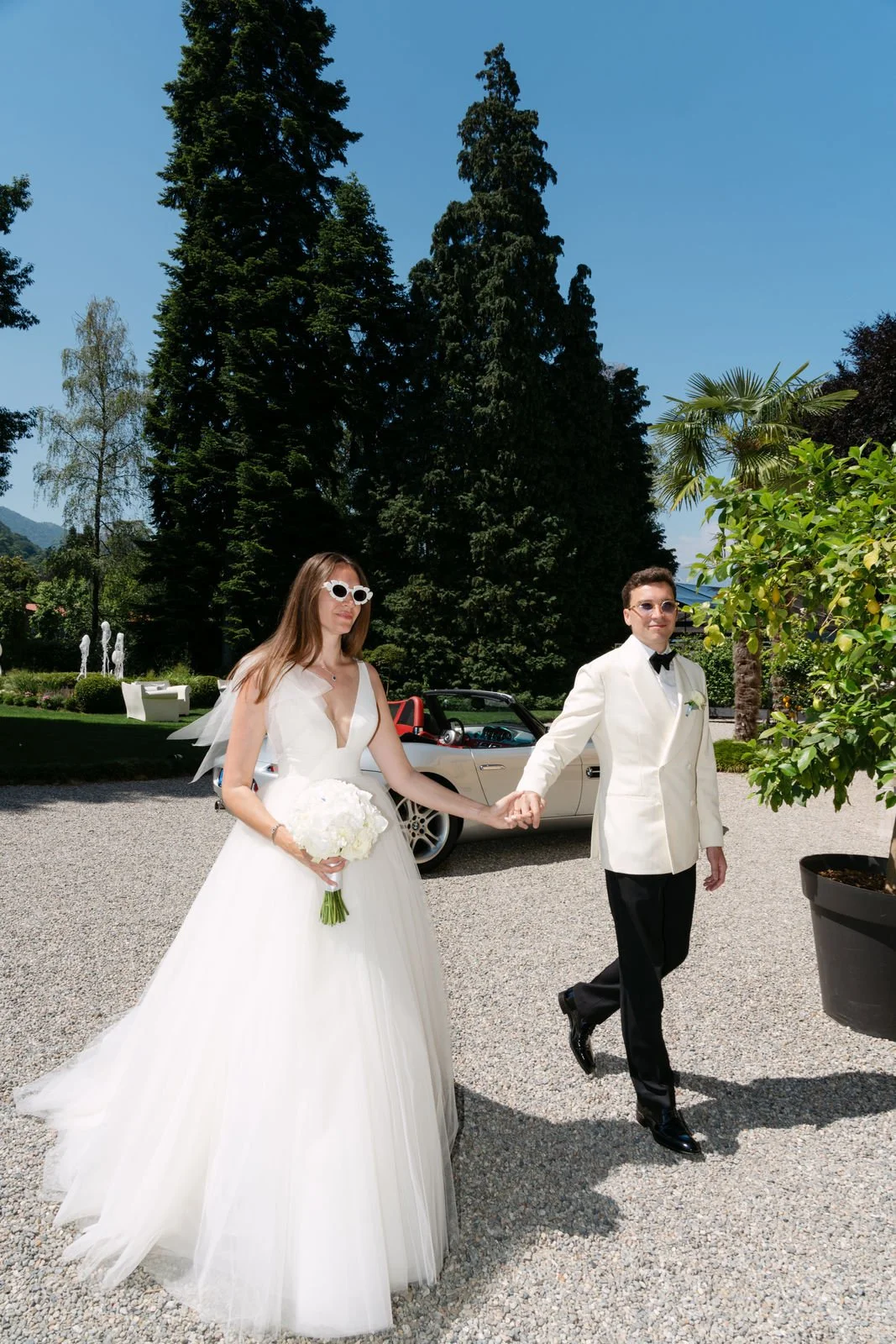 Ein Brautpaar beim Spaziergang im Garten, der Braut trägt ein weißes Hochzeitskleid und hält einen Blumenstrauß, der Bräutigam trägt eine weiße Frackjacke und schwarze Hosen, im Hintergrund ein rotes Auto und Grünpflanzen.