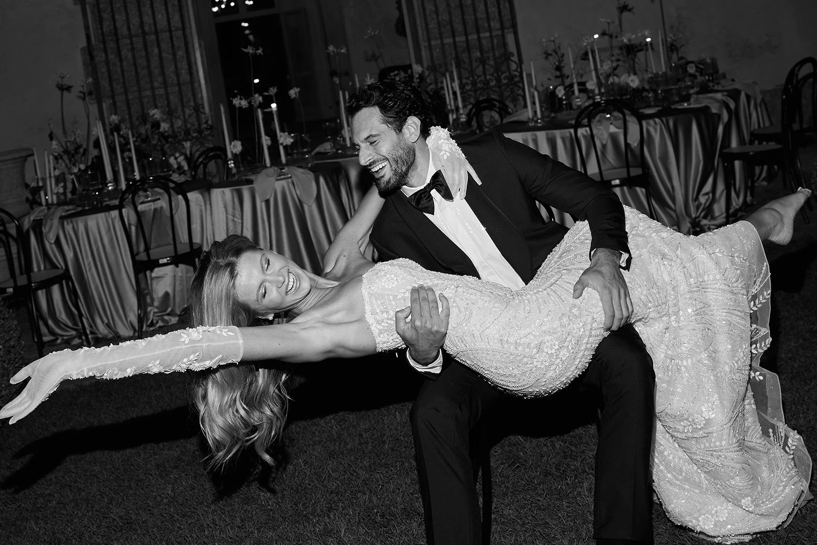 Ein lachendes Pärchen bei einer Hochzeitsfeier, der Mann trägt einen schwarzen Anzug mit Fliege, die Frau trägt ein elegantes Hochzeitskleid, sie tanzen in einem festlich gedeckten Saal.