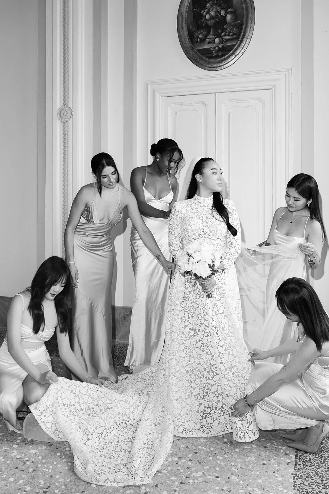 Eine Braut in einem weißen Spitzenkleid hält einen Brautstrauß, umgeben von fünf Frauen, die ihre Brautkleider zurechtrücken, in einem Raum mit weißen Wänden und einer verzierenden Tür.