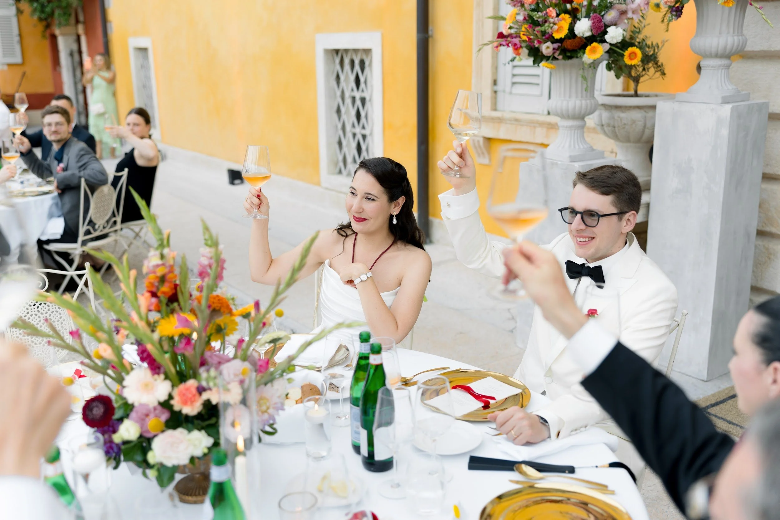 Menschen bei einer eleganten Feier oder Hochzeit, umgeben von Blumen und Tischdekorationen, die auf einer Terrasse oder im Freien sitzen und anstoßen.