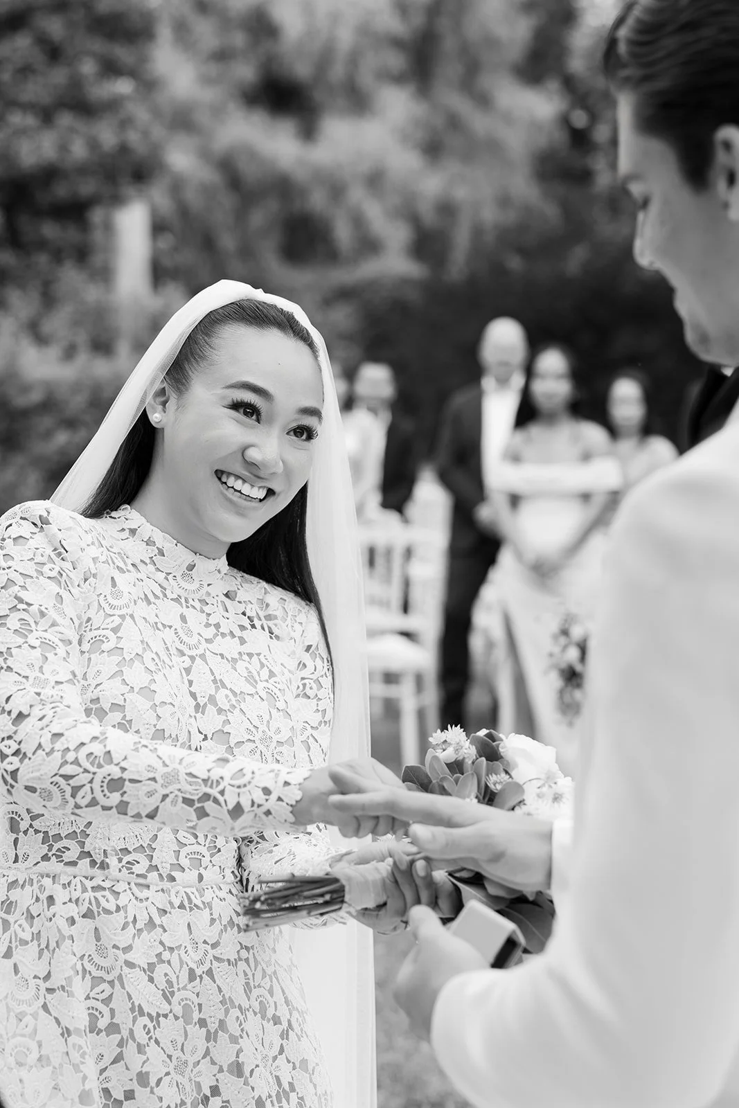 Braut und Bräutigam bei ihrer Hochzeit, die Braut trägt ein elegantes Spitzenkleid und hält Blumen, während sie sich die Hände reichen. Im Hintergrund sind Hochzeitsgäste im Freien, einige stehen, alle tragen formelle Kleidung.