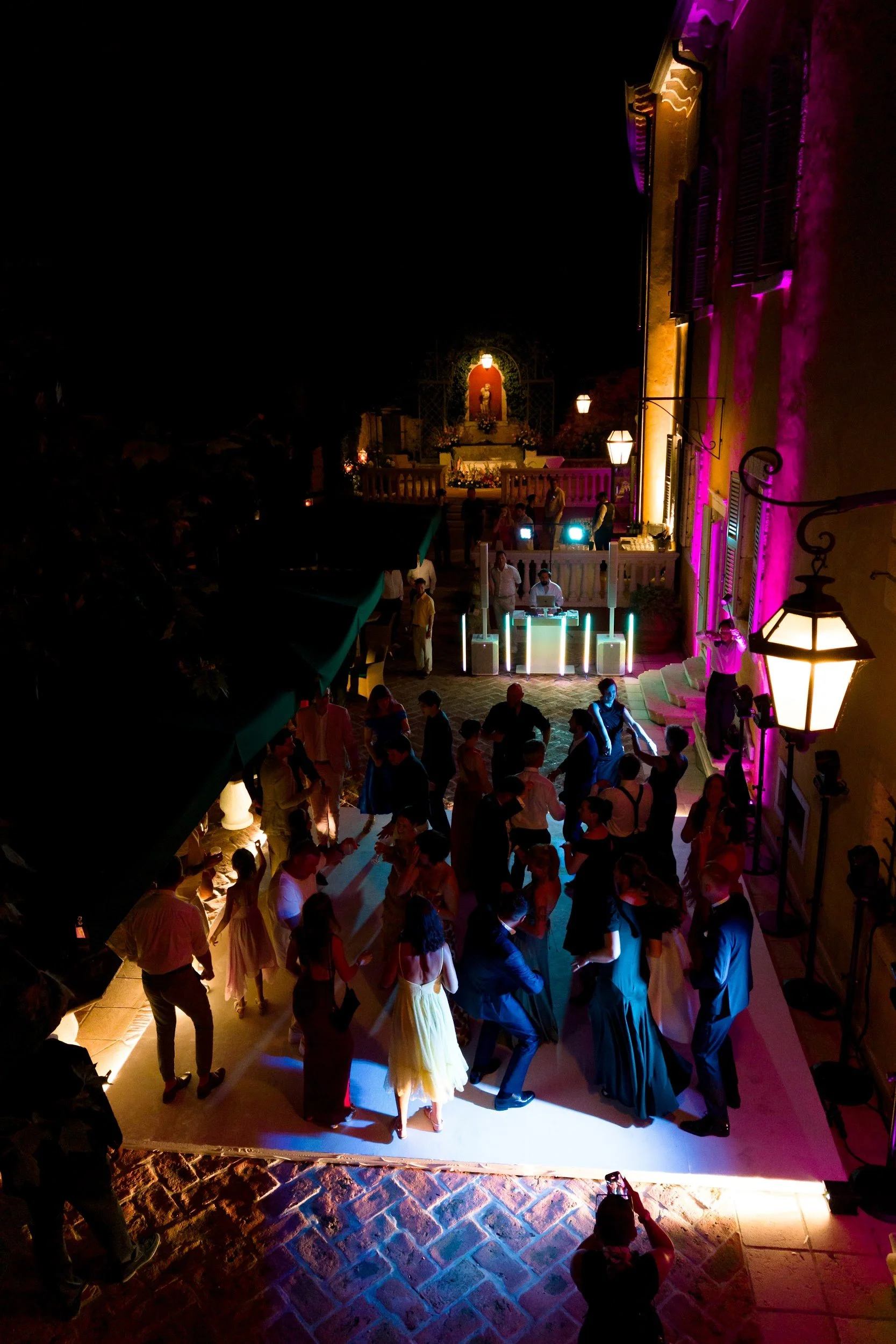 Eine nächtliche Szene mit einer Menschenmenge auf einem Tanzball im Freien, beleuchtet von bunten Lichtern, inklusive einer Begleitband und einem Altar im Hintergrund.
