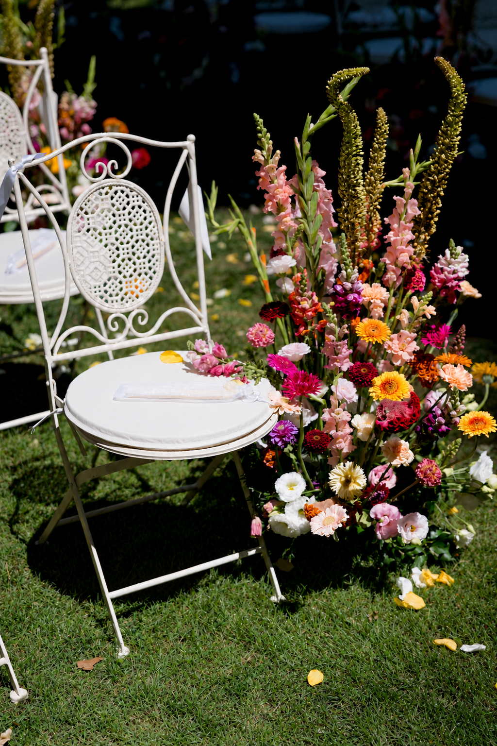 Ein weißer Gartenstuhl mit Kissen neben einem großen Blumenarrangement aus bunten Frühlingsblumen auf grünem Rasen bei sonnigem Wetter.