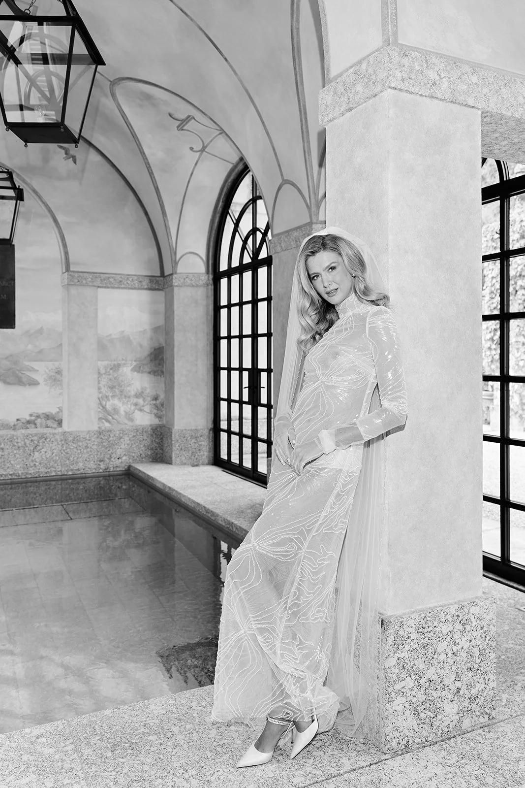 Schwarz-weiß-Bild einer jungen Frau in eleganter Hochzeitskleidung an einem Fenster in einem kunstvoll dekorierten Raum.