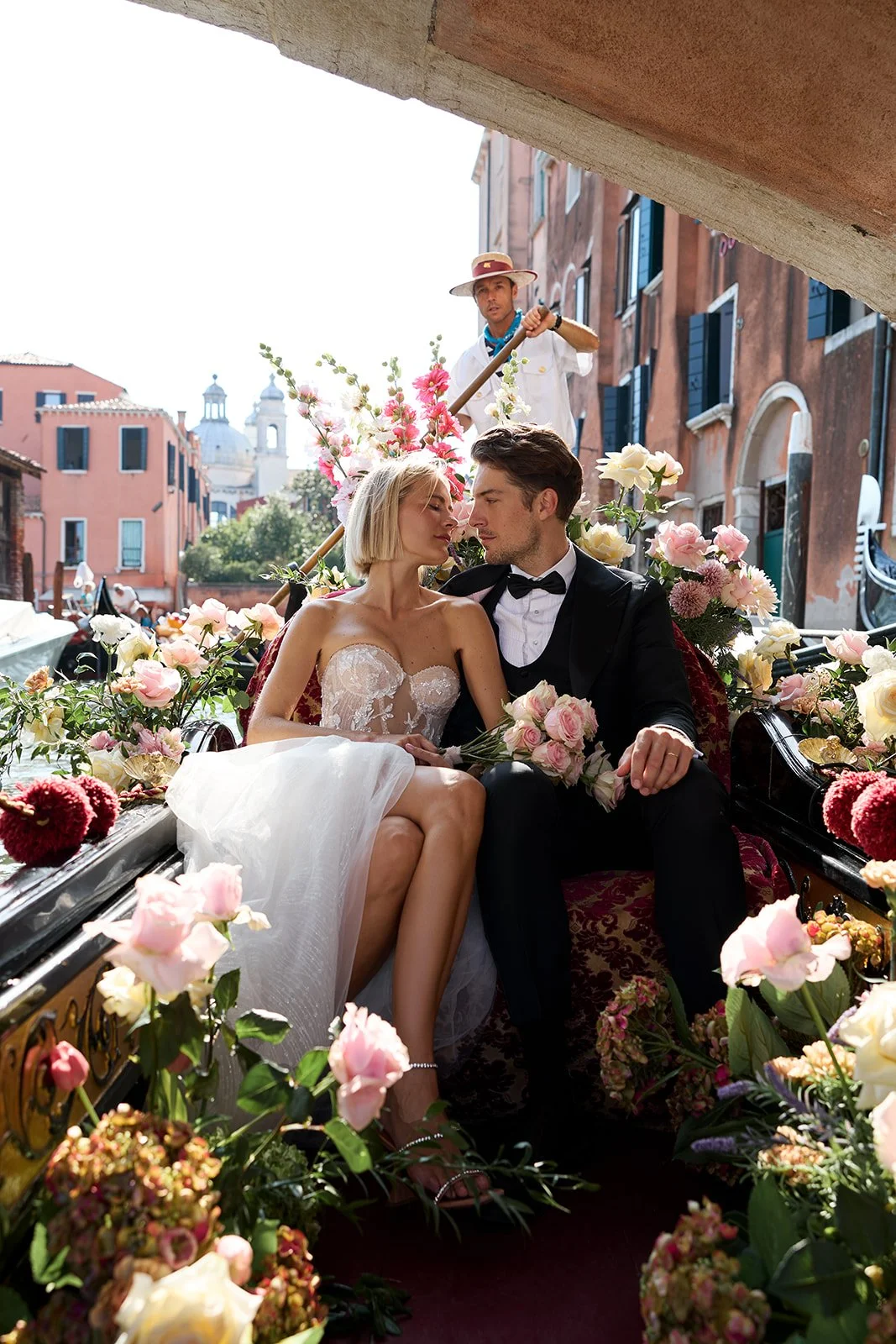 Ein Brautpaar sitzt auf einer mit Blumen geschmückten Kutsche in Venedig, umgeben von rosa und weißen Blumen, während eine Gondoliere im Hintergrund die Kutsche zieht.