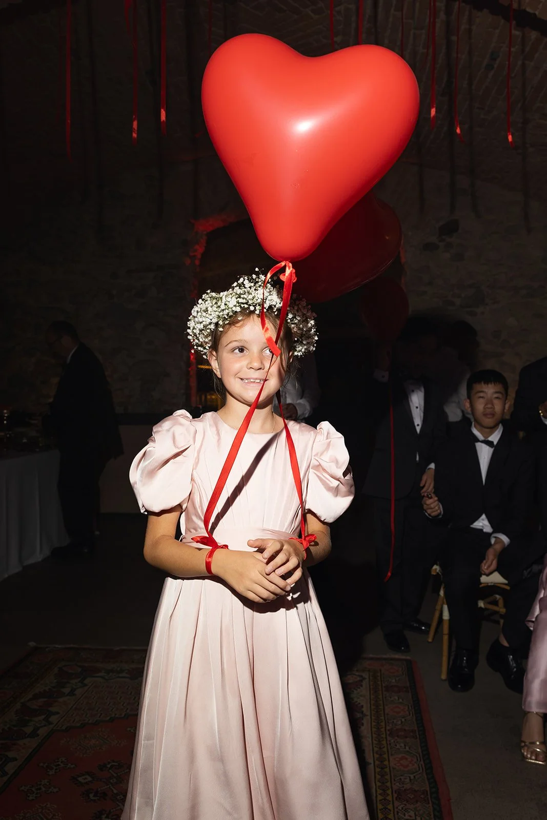 Ein Mädchen trägt ein rosa Kleid, eine Blumenkrone und hält ein Herz-förmigen Luftballon während einer Feier oder Hochzeit, umgeben von anderen Menschen in formeller Kleidung.