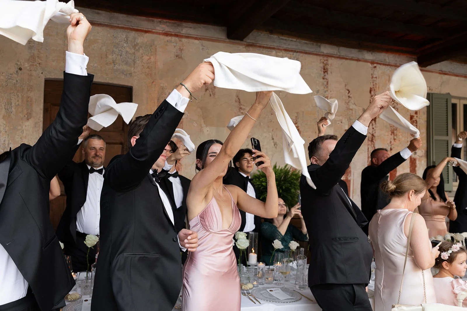 Menschen, die bei einer Feier weiße Tücher in die Luft werfen, vermutlich bei einer Hochzeit, in einem rustikalen Raum mit Steinwänden.