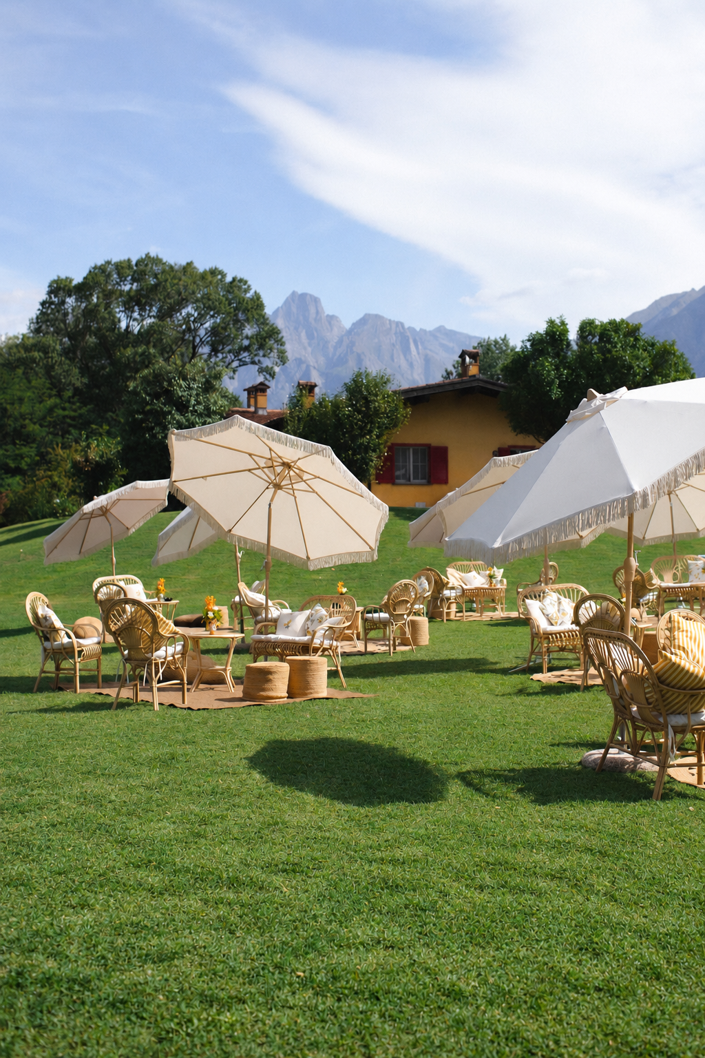 Ein Garten mit mehreren weißen Sonnenschirmen, Holzstühlen und Tischen auf grünem Rasen, im Hintergrund ein gelbes Haus mit roten Fensterläden, umgeben von Bäumen und Bergen im Gebirge.