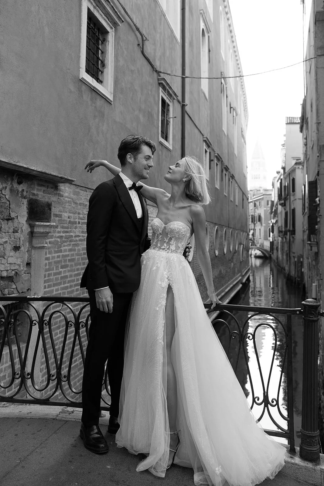 Ein frisch vermähltes Paar in formeller Kleidung steht an einer Brüstung an einem Kanal in Venedig, Italien, im schwarzen und weißen Foto.