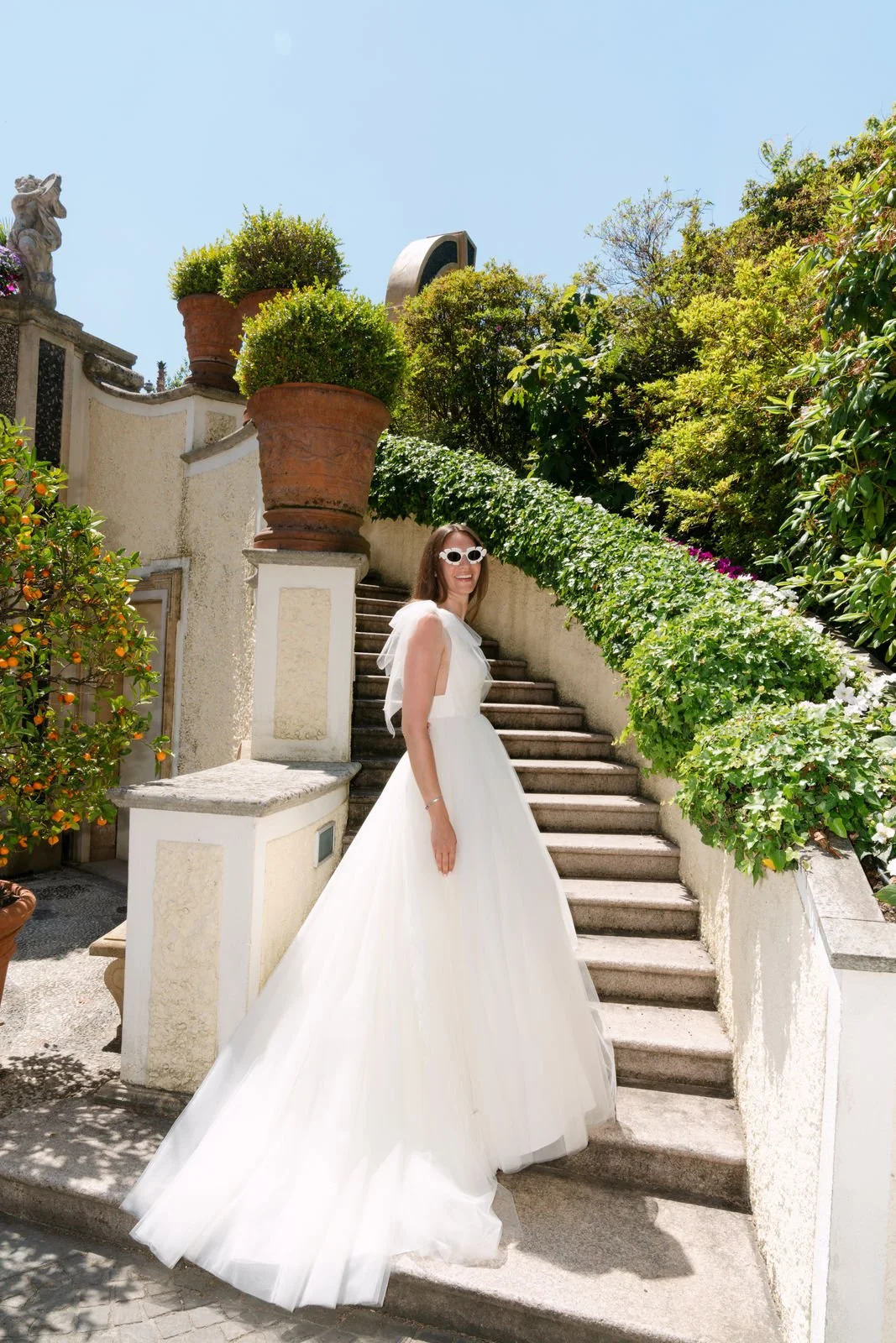 Frau in weißem Brautkleid mit großen Sonnenbrillen, die auf einer Treppe bei sonnigem Wetter steht, umgeben von grünen Pflanzen und Terrakotta-Tontöpfen.