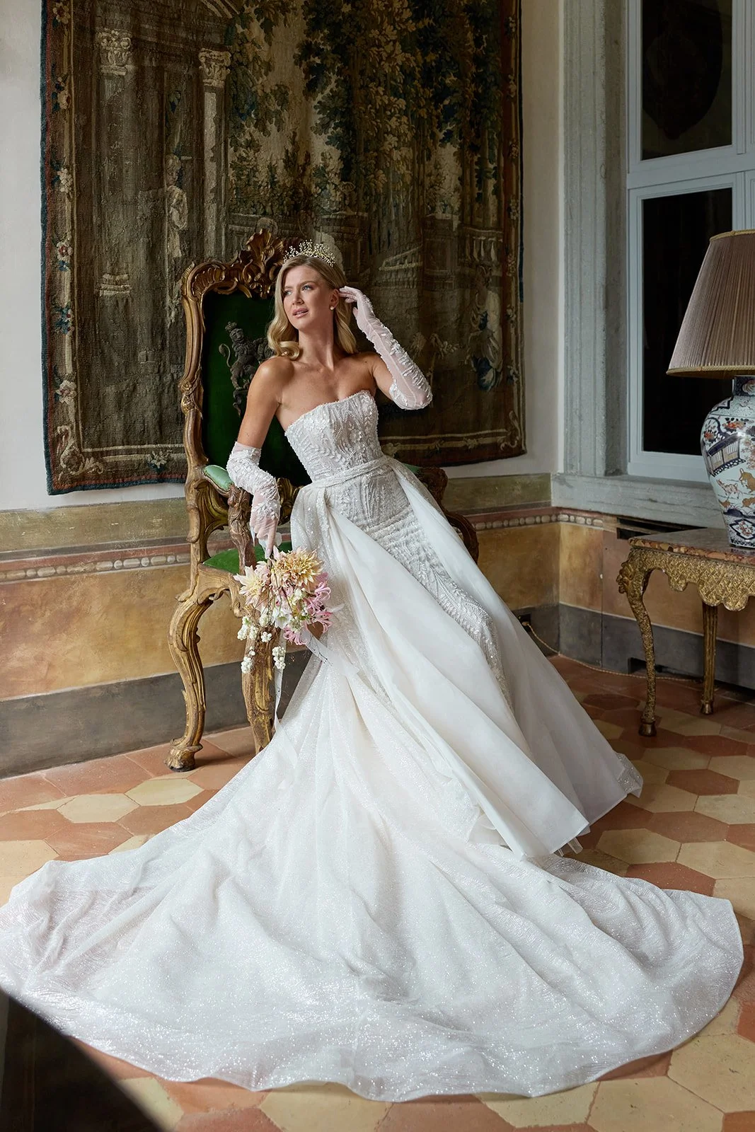 Eine Frau in einem weißen Brautkleid sitzt auf einem antiken, grün gepolsterten Stuhl im Raum, trägt eine Tiara, hält einen Blumenstrauß und schaut nach oben. Im Hintergrund hängt ein Wandteppich, und es sind ein großer Lampenschirm, eine antike Komm