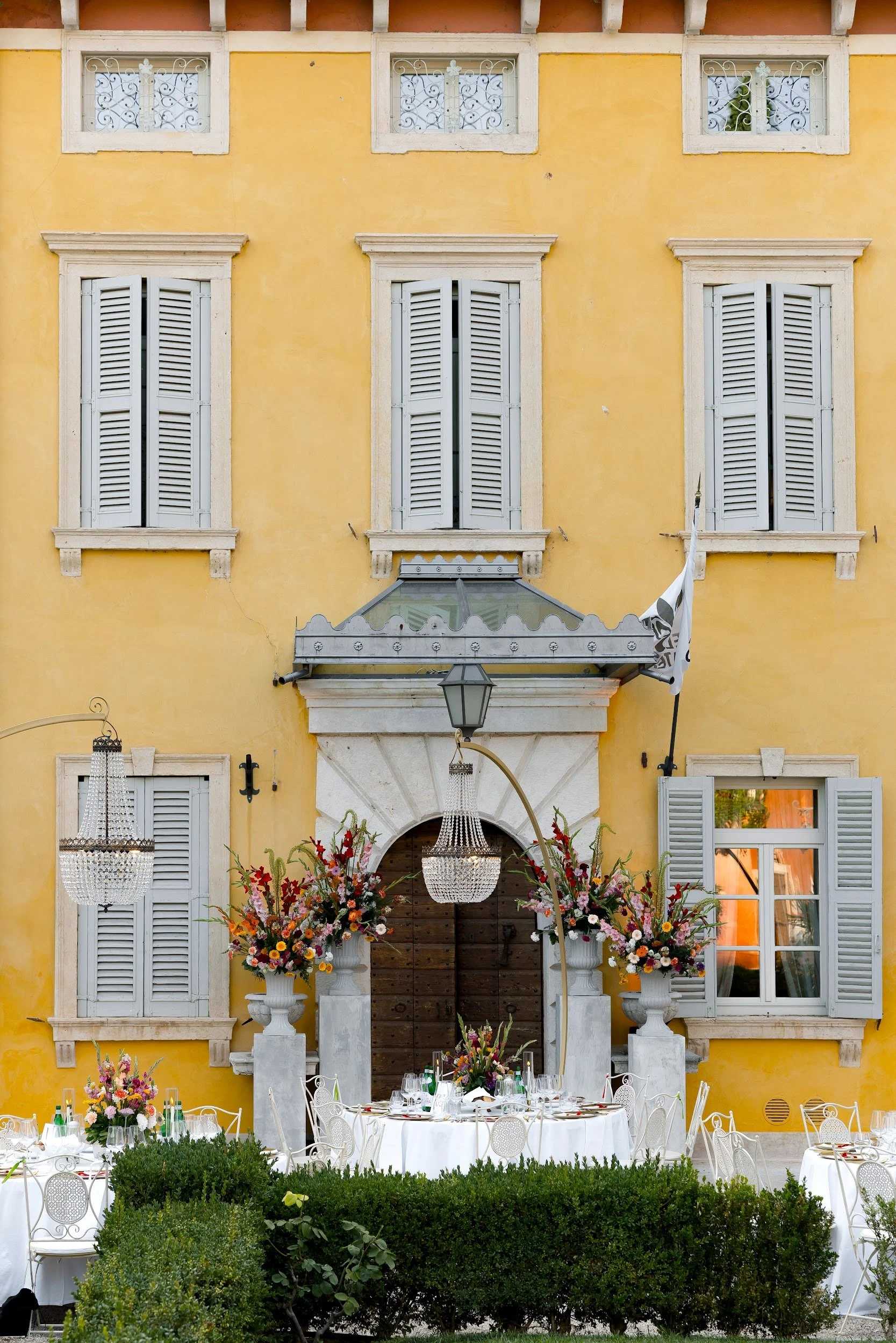 Ein festlich gedeckter Tisch im Freien vor einem gelben Haus mit weißen Fensterläden. Der Tisch ist mit Blumenarrangements, Kerzen und Gläsern geschmückt, und mehrere Kronleuchter hängen darunter, um eine elegante Atmosphäre zu schaffen.