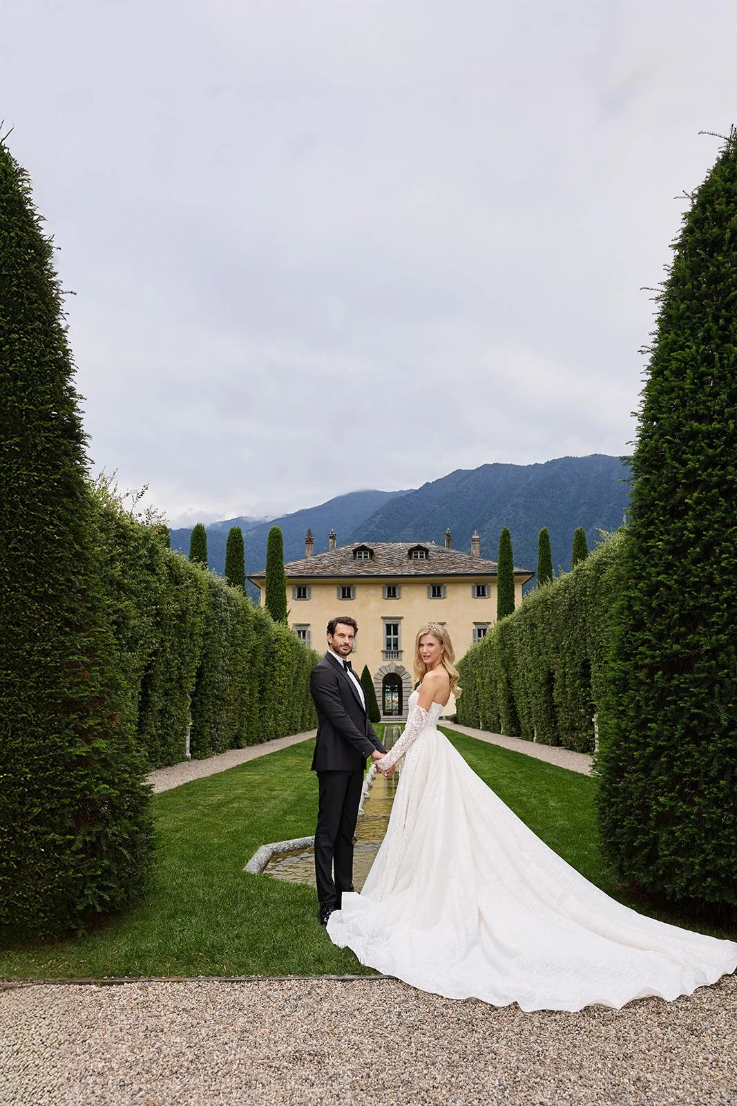 Ein Brautpaar in Hochzeitskleidung, hält Händchen vor einem großen, eleganten Herrenhaus, umgeben von gepflegten Gärten und hohen Hecken, im Hintergrund Berge und bewölkter Himmel.