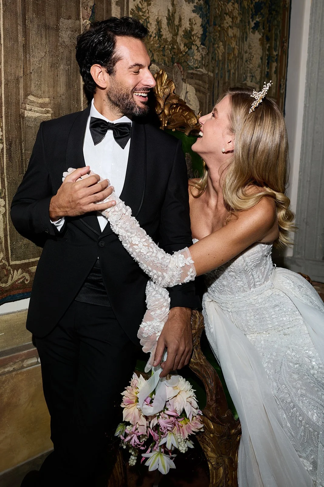 Ein Brautpaar bei ihrer Hochzeit. Der Bräutigam trägt einen schwarzen Anzug mit Fliege, die Braut trägt ein weißes Brautkleid mit Spitze und eine kleine Krone im Haar. Sie lachen sich an.