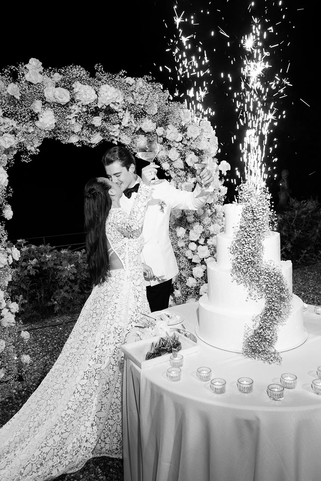 Ein Paar bei einer Hochzeit, das mit Sektgläsern anstößt, vor einer mehrstöckigen Hochzeitstorte mit Feuerwerk, umgeben von Blumen, im Freien bei Nacht.