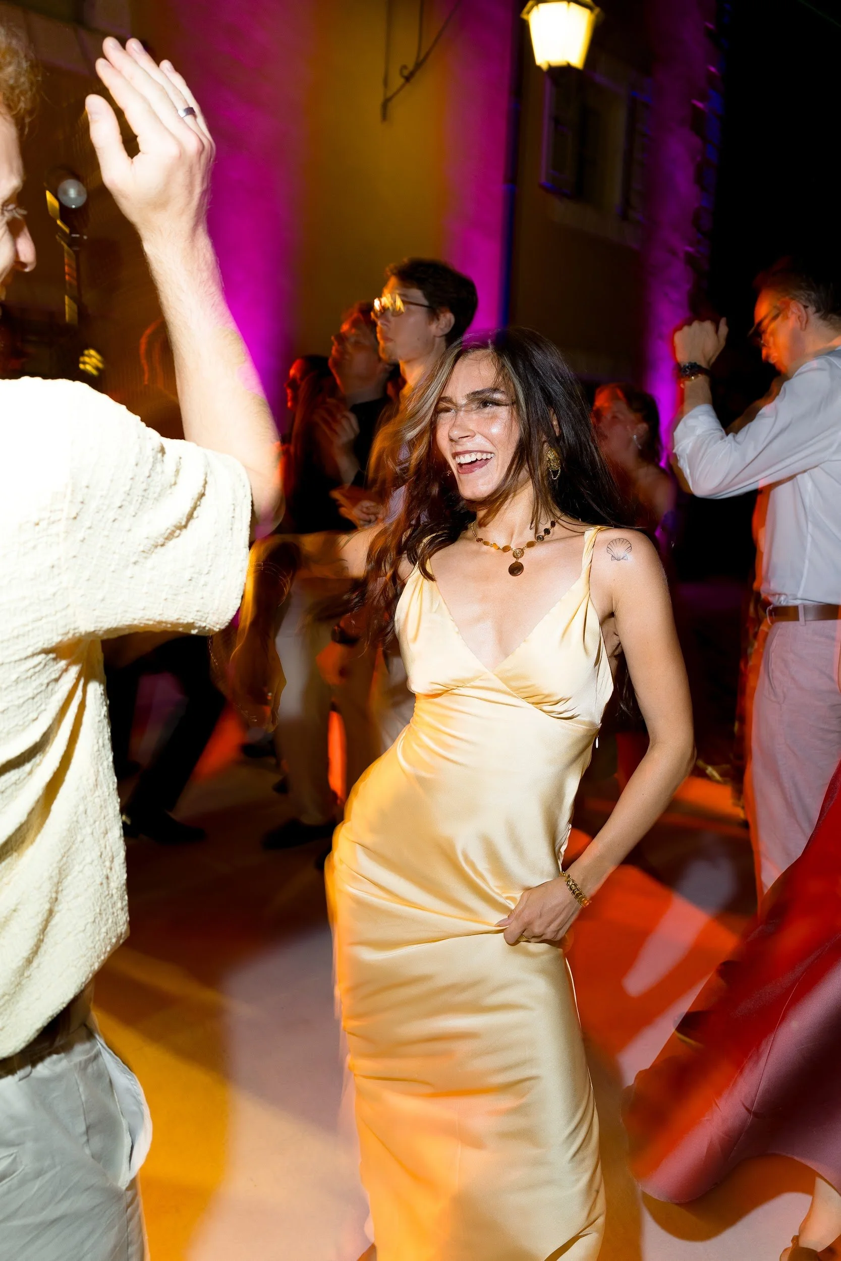 Eine fröhliche Frau in einem goldgelben Kleid tanzt auf einer Party, umgeben von anderen tanzenden Personen, während bunte Lichter die Szene beleuchten.