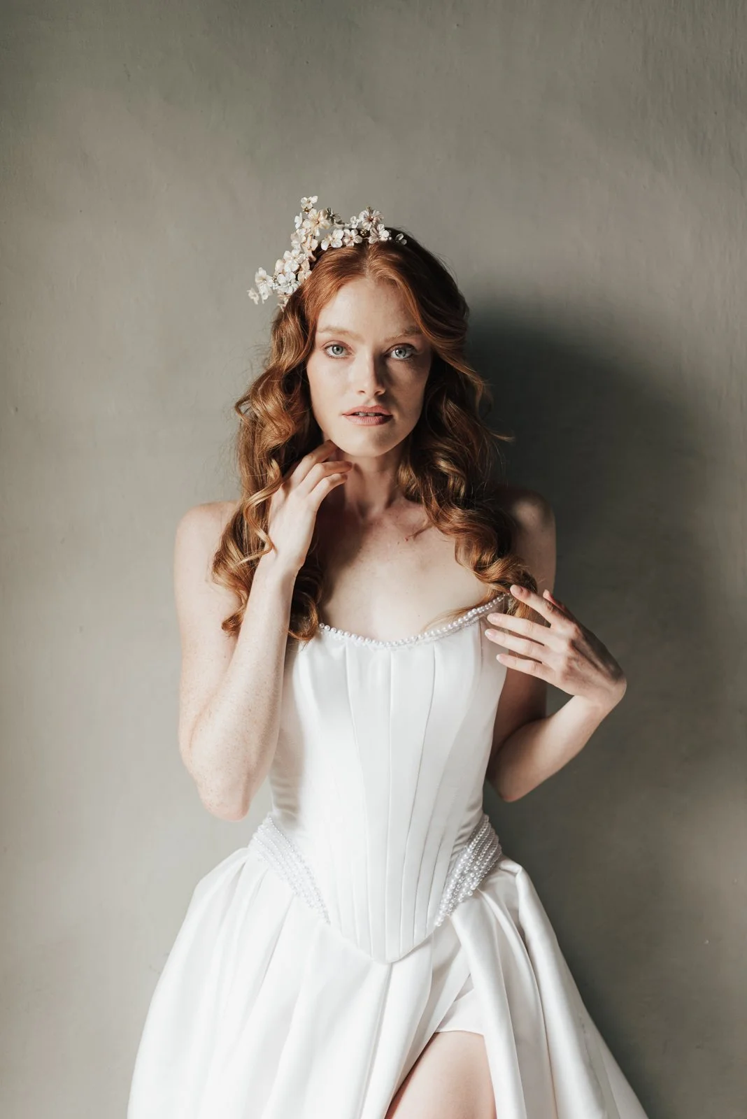 Frau in einem weißen Kleid mit Blumen im Haar posed vor einer Wand.