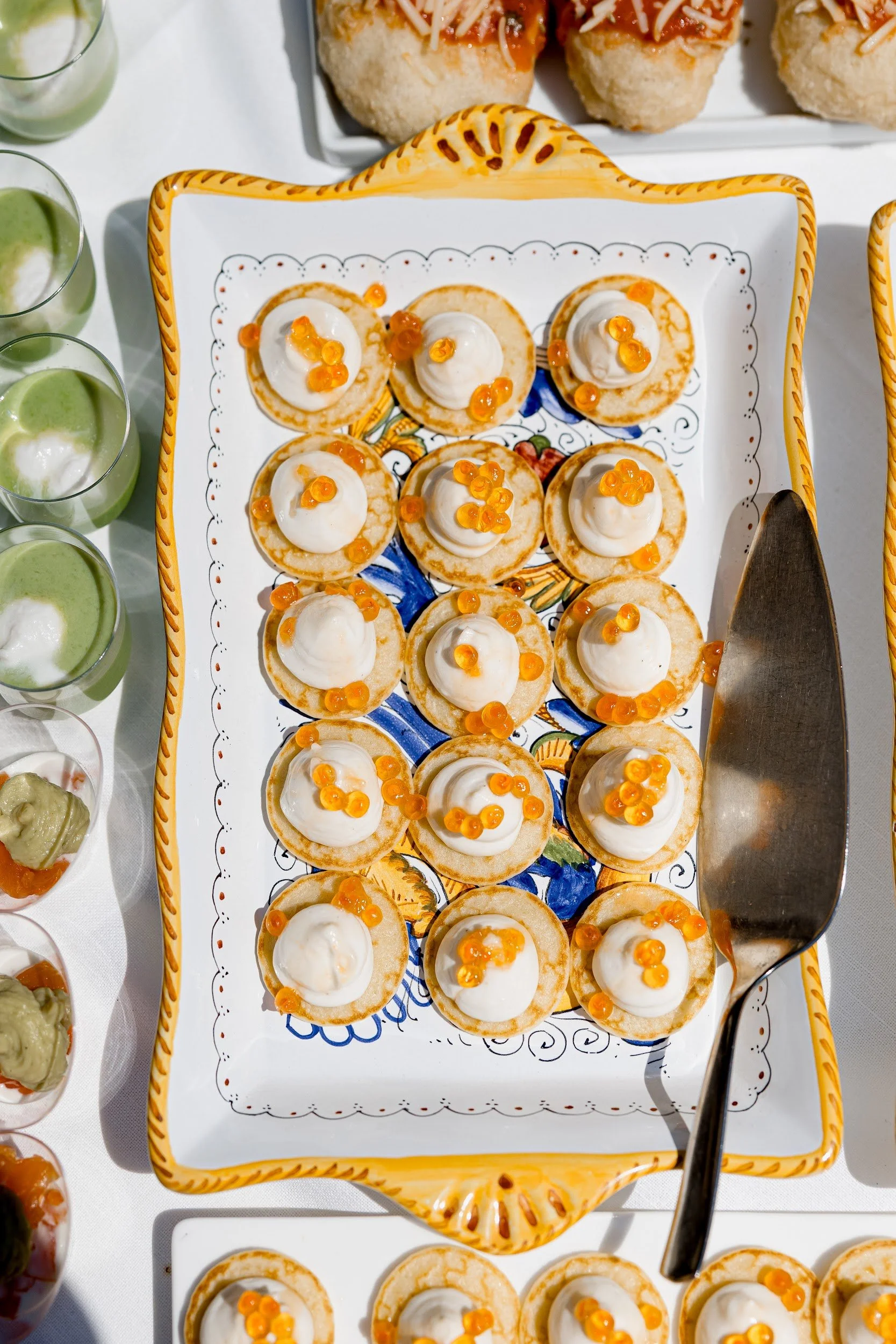 Dessertplatte mit Törtchen, die mit Sahne und orangefarbenen Beeren garniert sind, auf einer dekorativen Keramikplatte, umgeben von kleinen Aufsätzen mit grünem und anderem Dip.