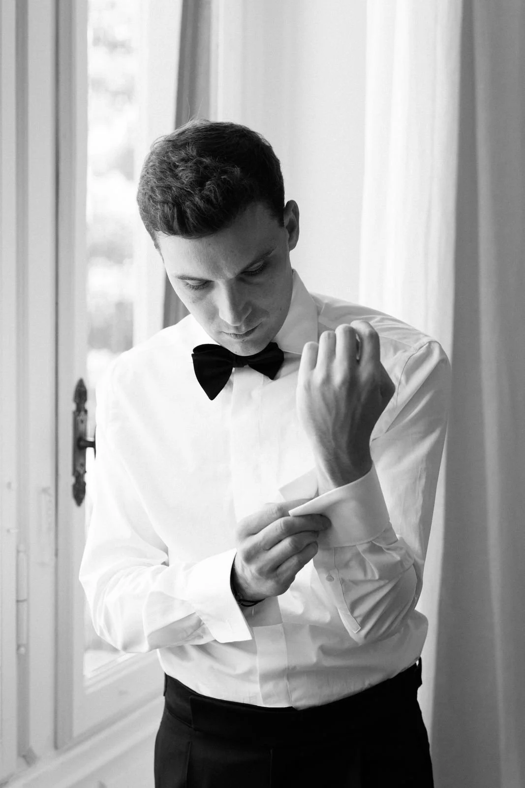 Junger Mann in formeller Kleidung mit Fliege beim Anziehen eines Hemdmanschettenknopfs, schwarz-weiß Aufnahme, im Hintergrund Fenster und Vorhänge.