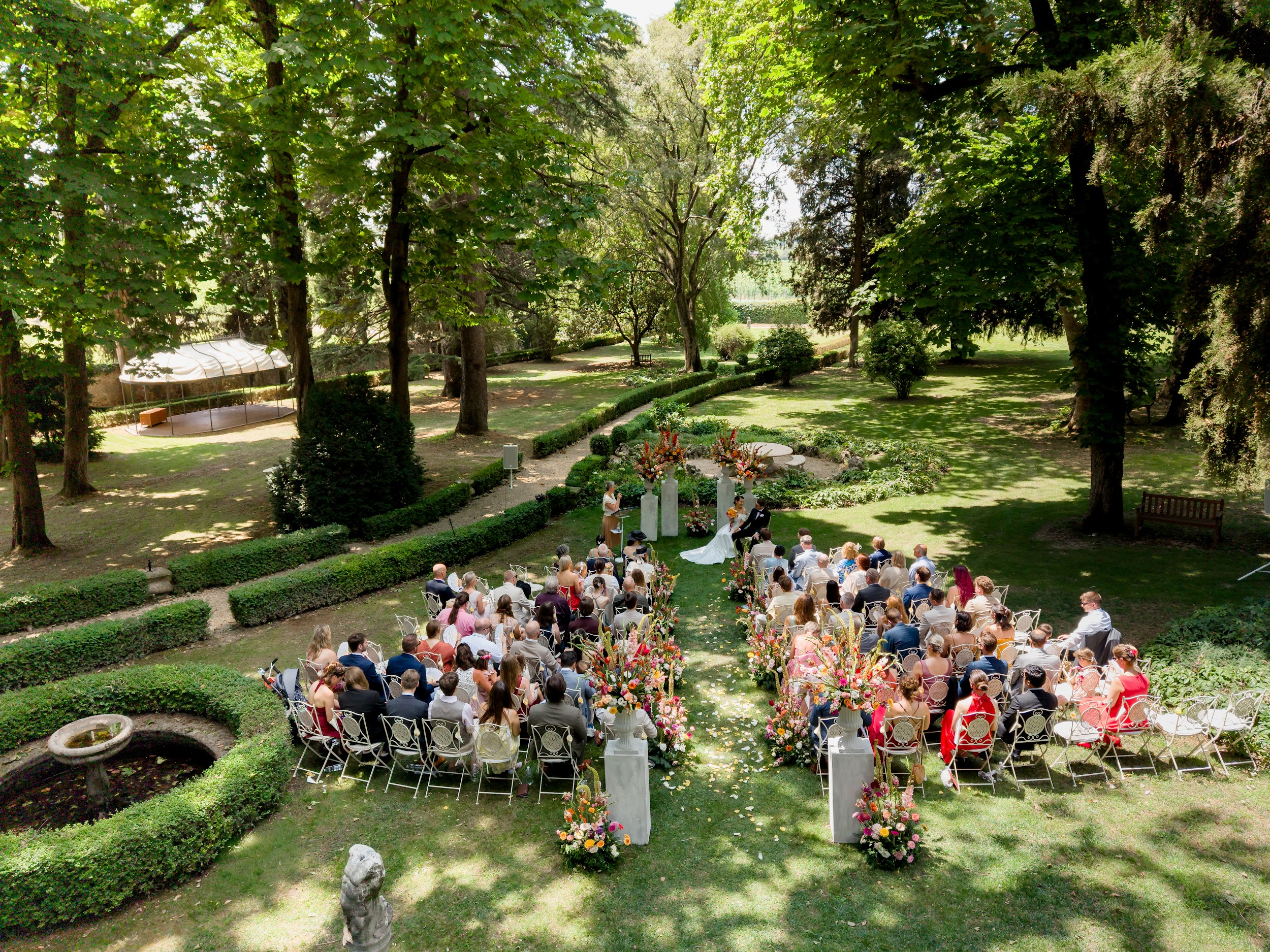 Eine Hochzeitszeremonie im Freien in einem Garten mit vielen Zuschauern, grünen Bäumen und bunten Blumen, ein Brautpaar bei der Trauung, andere Gäste sitzen und beobachten.