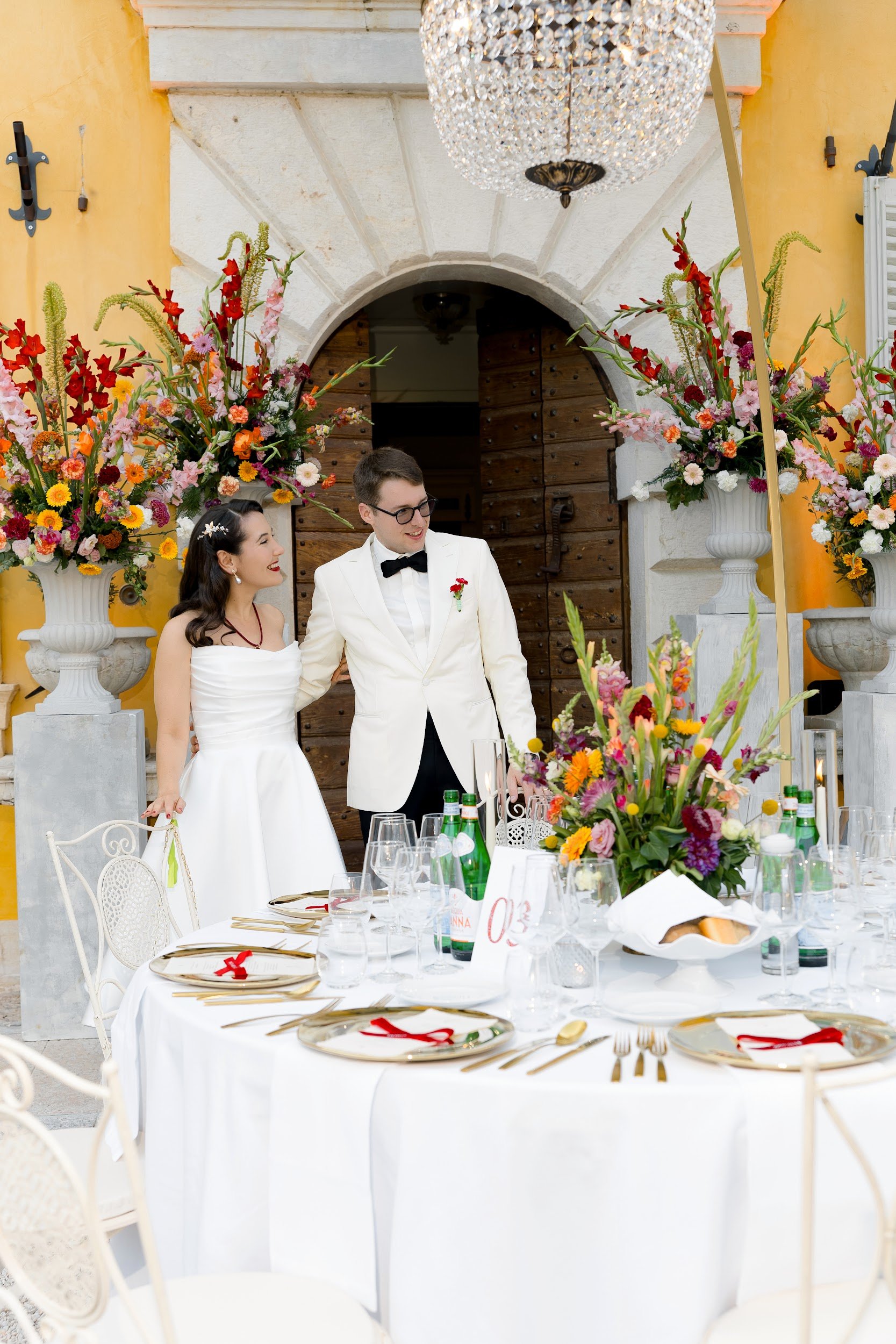 Ein Hochzeitspaar, ein Mann in weißem Anzug mit Fliege und eine Frau in einem weißen Brautkleid, stehen vor einem festlich gedeckten Tisch mit weißen Tischtüchern, goldenen Servierten und leuchtenden Blumenarrangements, im Hintergrund eine offene Hol