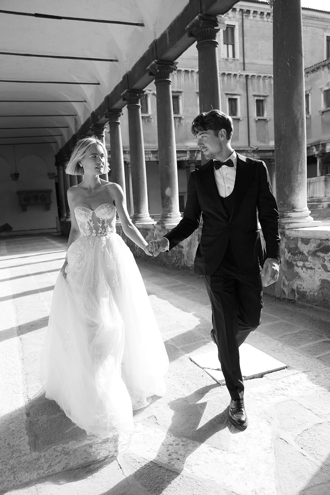 Ein schwarz-weiß Foto eines Paares bei einer Hochzeit, das sich an einem historischen Gebäude hält, die Frau trägt ein wunderschönes Hochzeitskleid, der Mann ein Anzug mit Fliege.