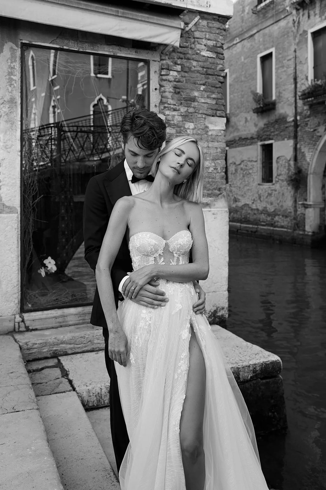 Ein Paar in eleganter Hochzeitskleidung steht am Wasser in einer alten Stadt mit Backstein-Gebäuden, im Schwarz-Weiß-Stil fotografiert.